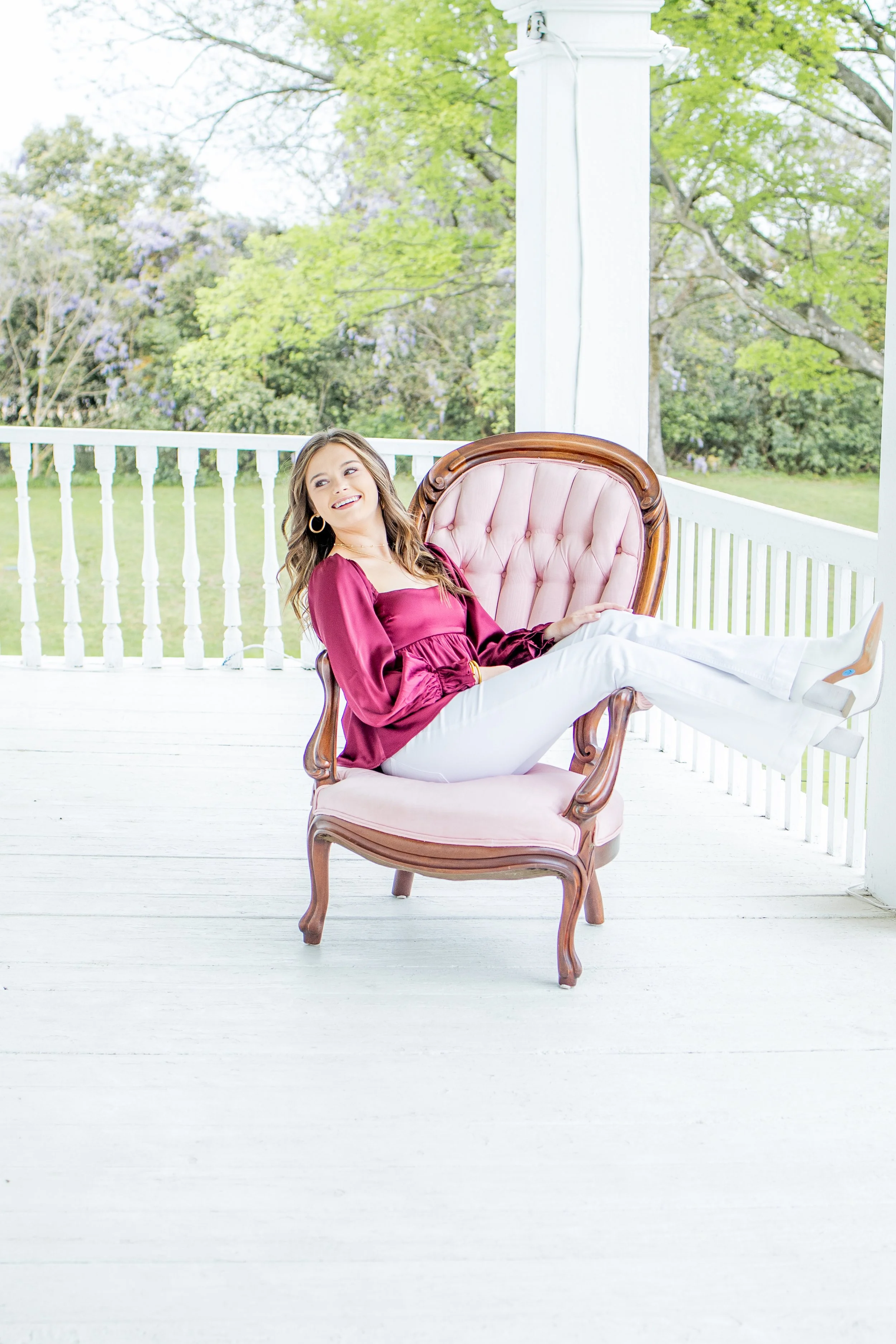 Young woman with long hair sitting on a vintage pink upholstered armchair on a white porch, wearing a maroon blouse and white pants, smiling with a background of green trees and partly cloudy sky.