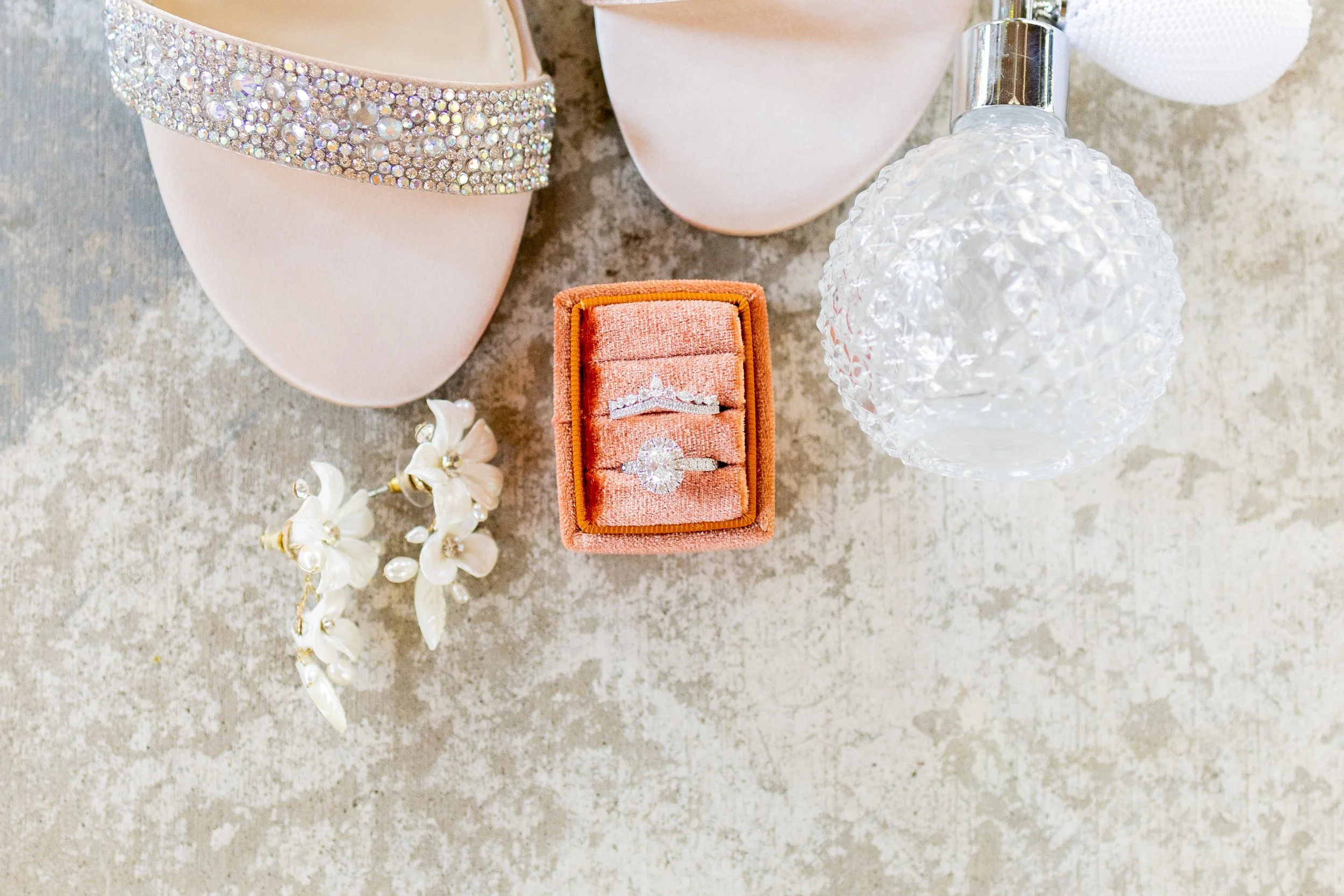 Close-up of wedding accessories including a pair of pink satin shoes adorned with rhinestones, floral earrings with pearls, a small box with diamond rings, a decorative perfume bottle, and wearing jewelry, all on a light-colored surface.