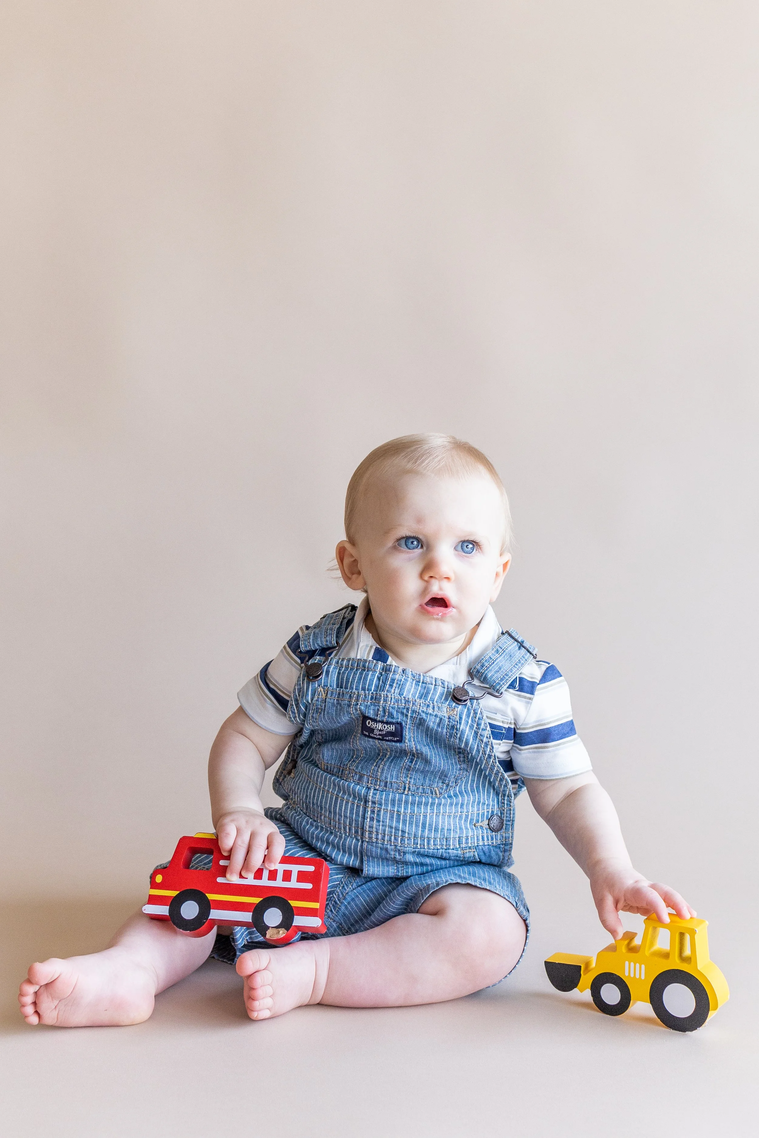 Young child with blue eyes sitting on the floor, holding a toy fire truck in one hand and a toy construction vehicle in the other, wearing a striped shirt and denim overalls.