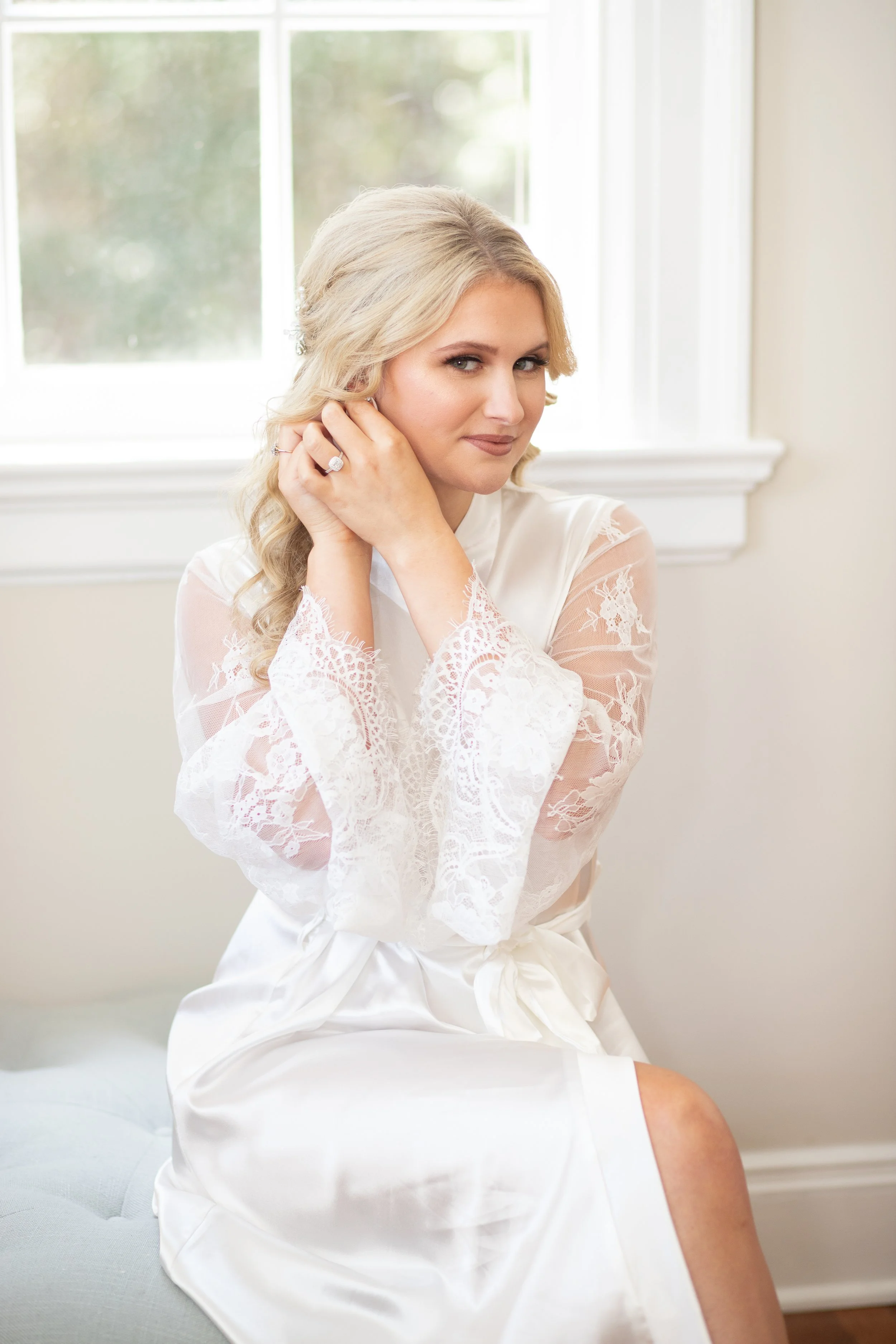 A woman with blonde hair, wearing a white lace robe, sitting by a window, adjusting an earring.
