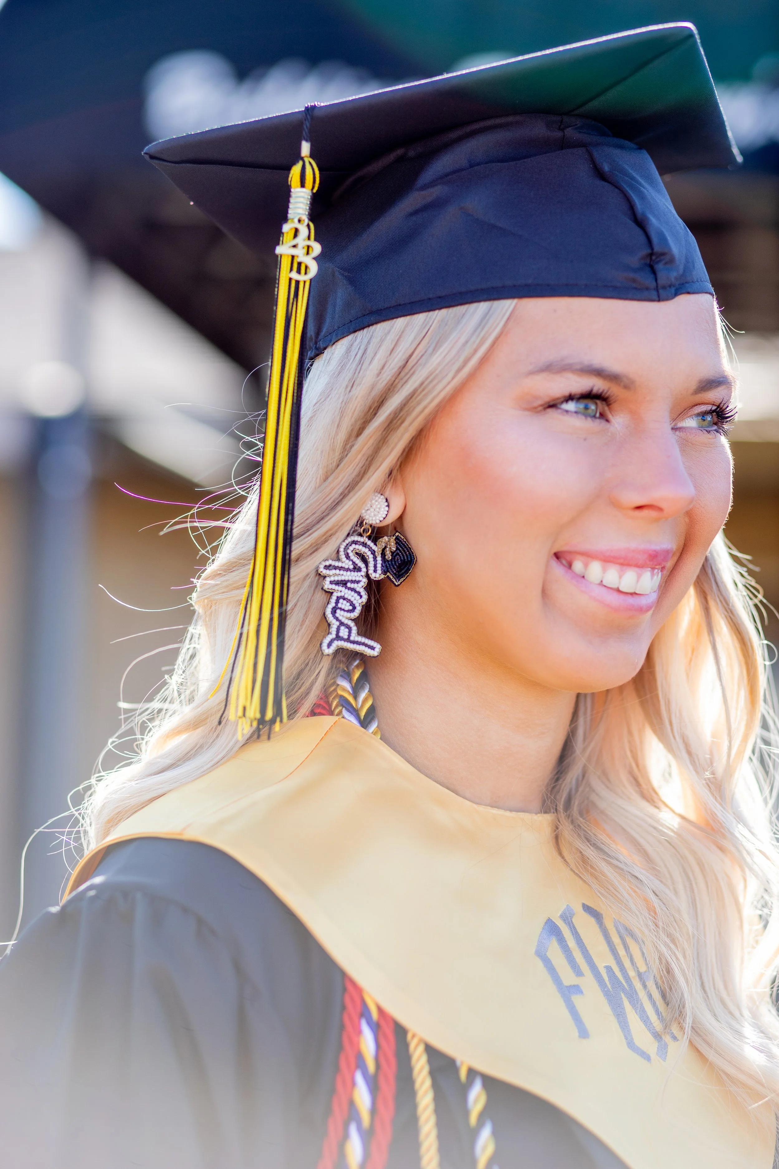 A young woman in graduation cap and gown smiling outdoors, wearing colorful honor cords and a decorative earring.