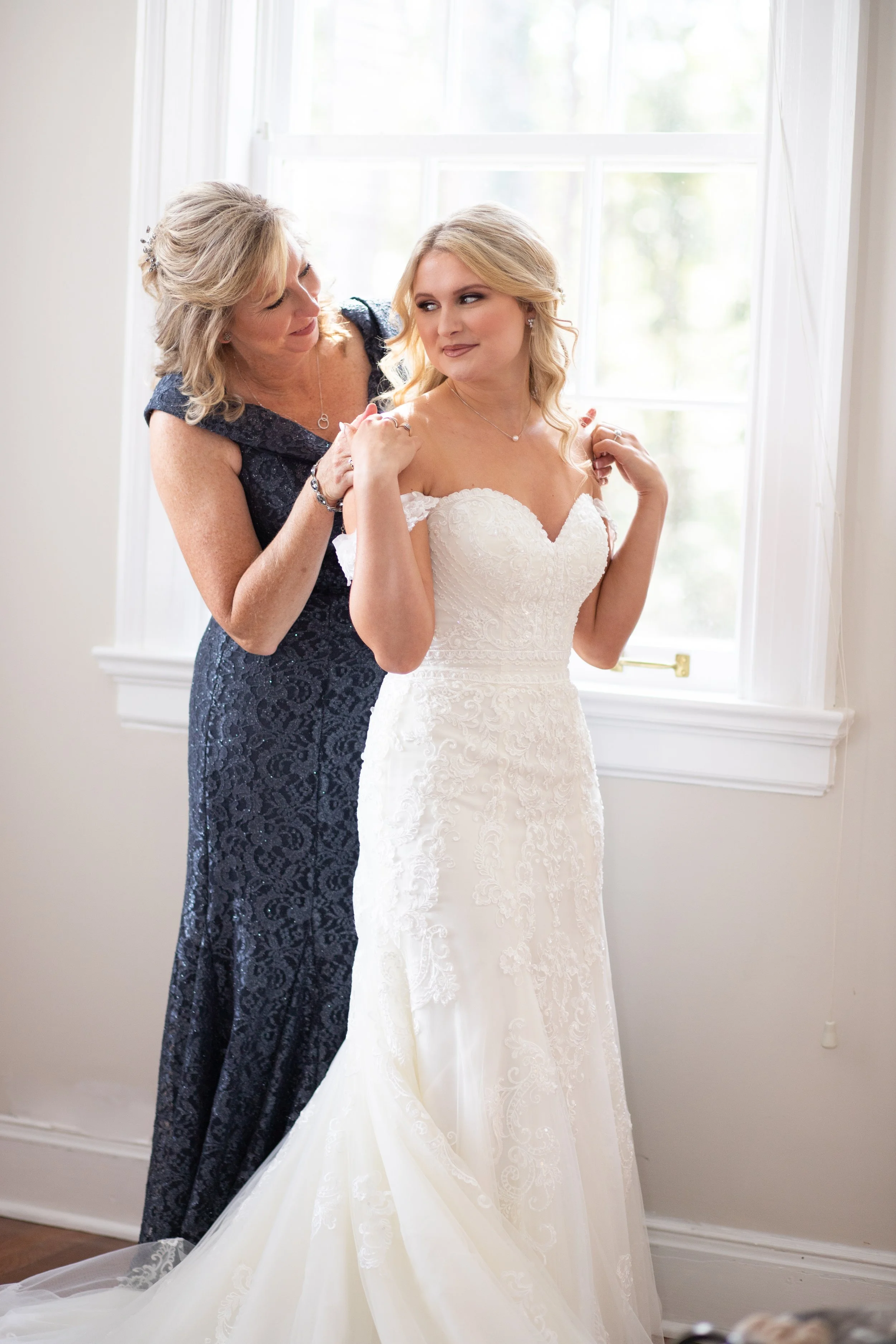 A bride in a white wedding dress is being helped by a woman in a dark blue lace dress, possibly her mother, as she prepares for her wedding. They are standing near a window, with sunlight shining in.