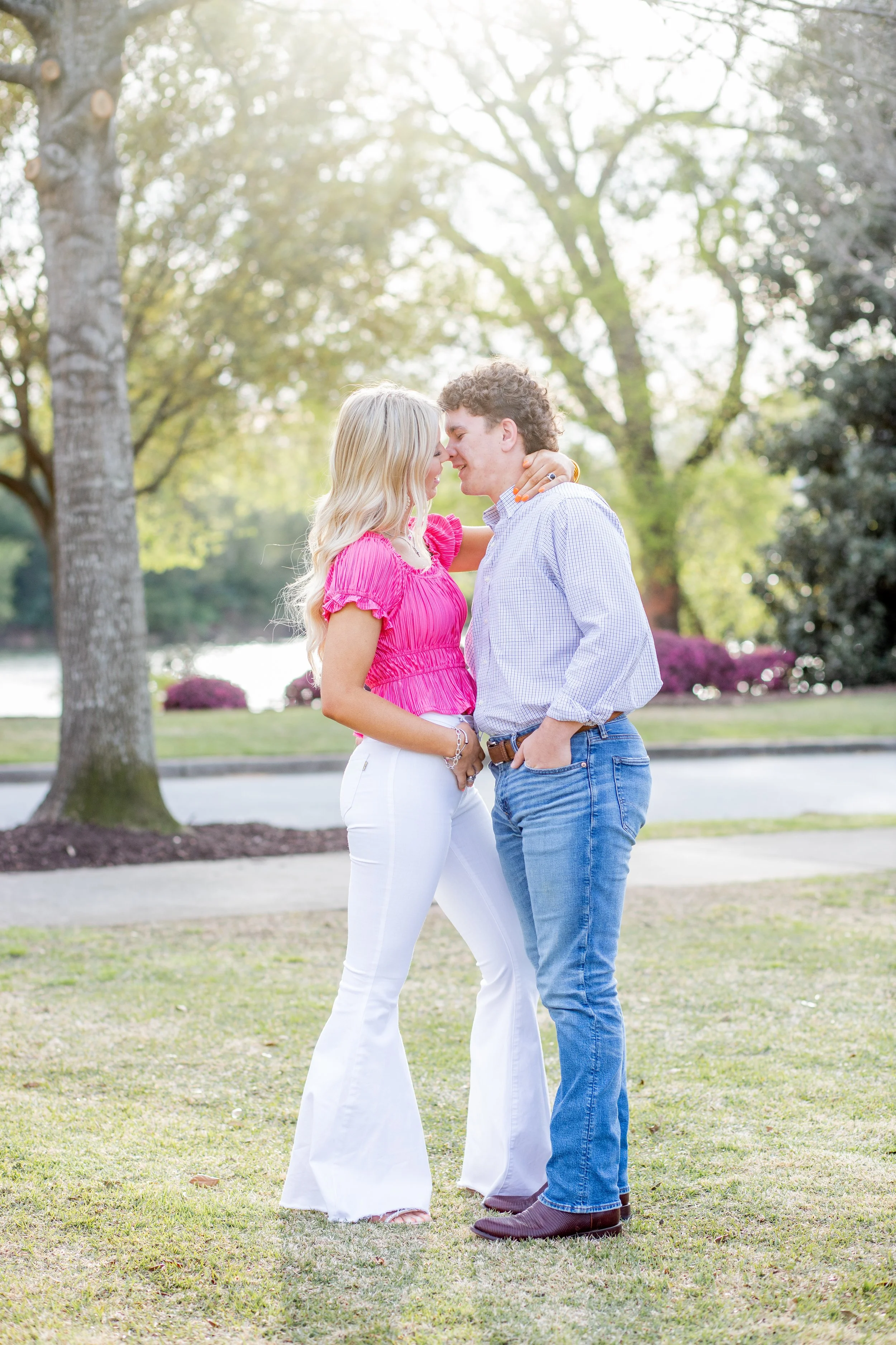 A young couple stands close together outdoors in a park, about to kiss, with trees and a river in the background, during daytime.