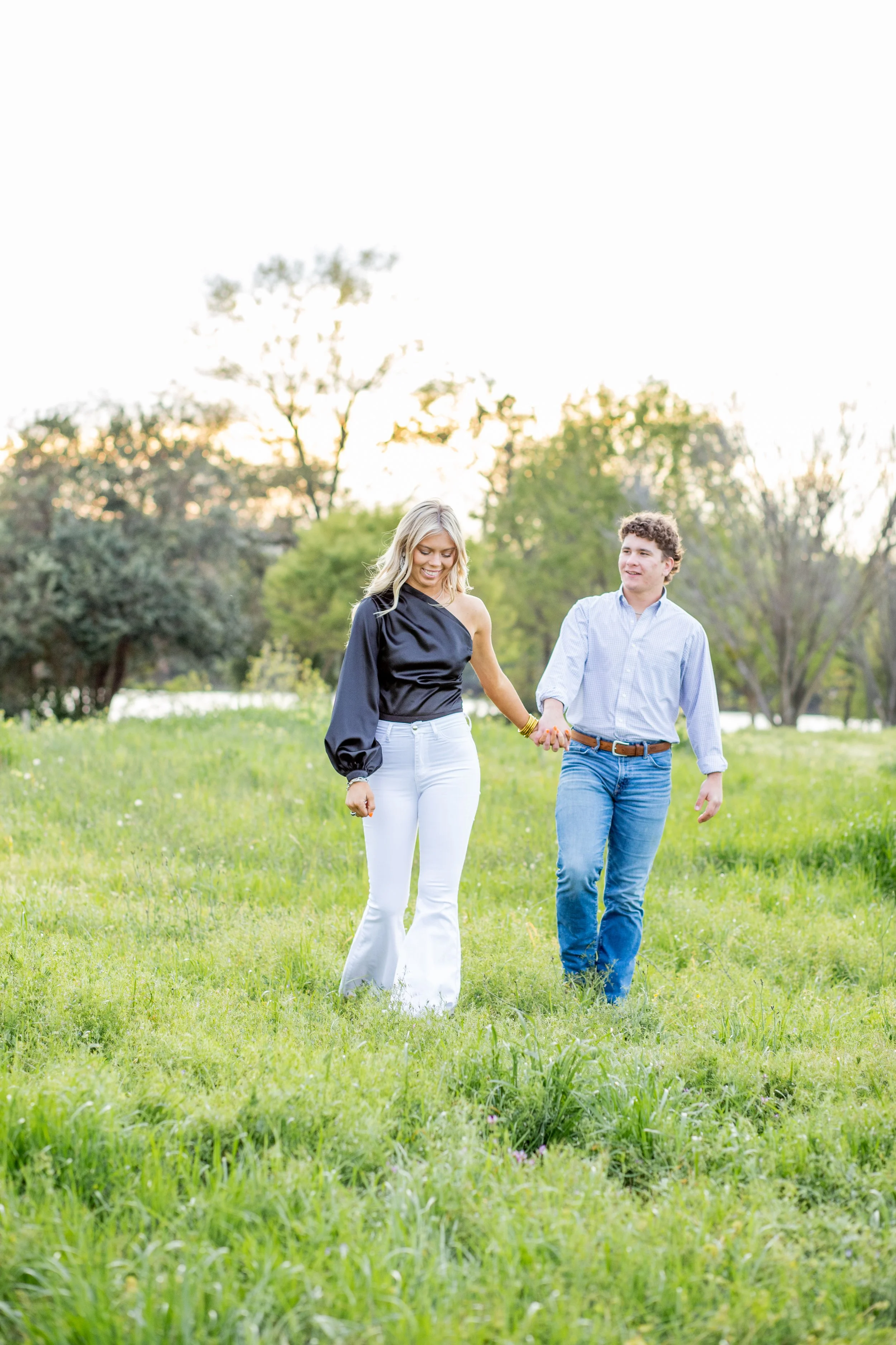 A couple walking hand-in-hand in a grassy park with trees in the background during sunset.