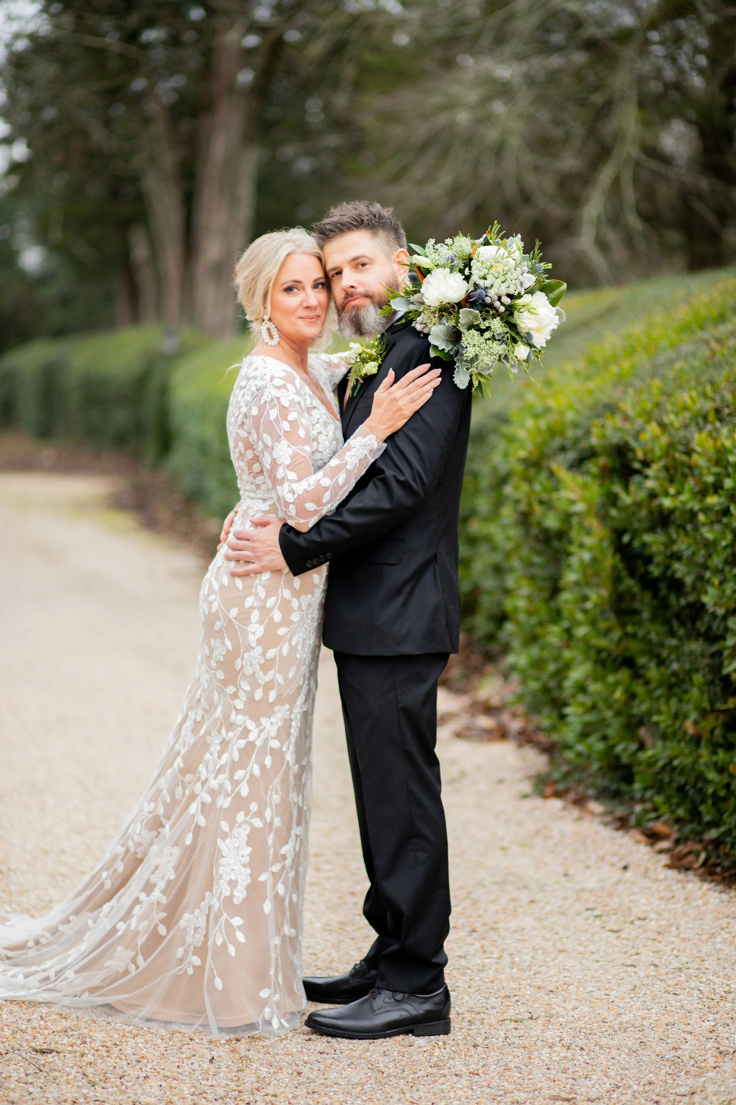 A bride and groom stand close together outdoors surrounded by greenery, with her holding a bouquet of white and green flowers over his shoulder while smiling at the camera.
