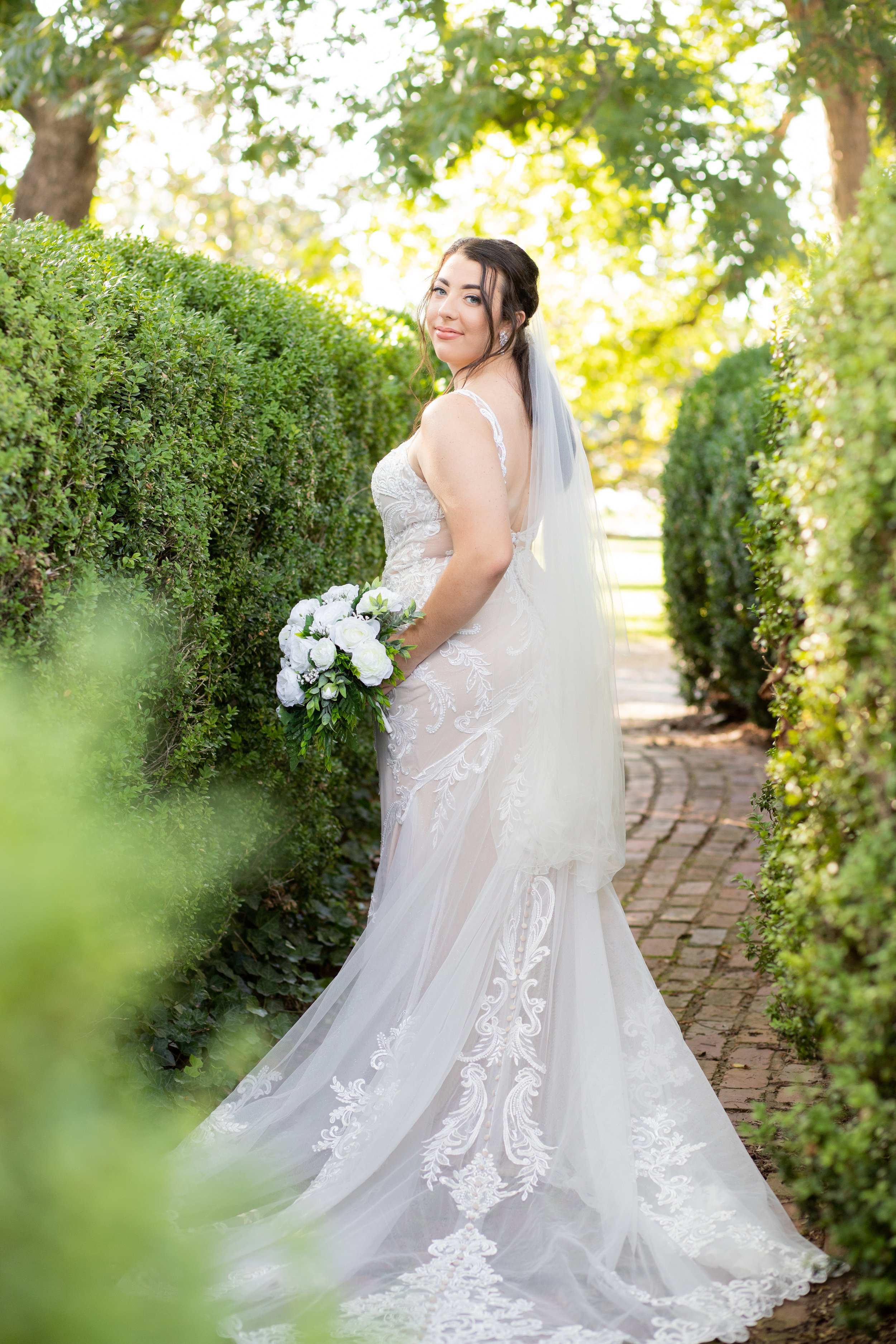 A bride in a white lace wedding dress stands on a brick pathway between green hedges, holding a bouquet of white flowers. She has dark hair and is wearing a long veil, with trees and sunlight in the background.
