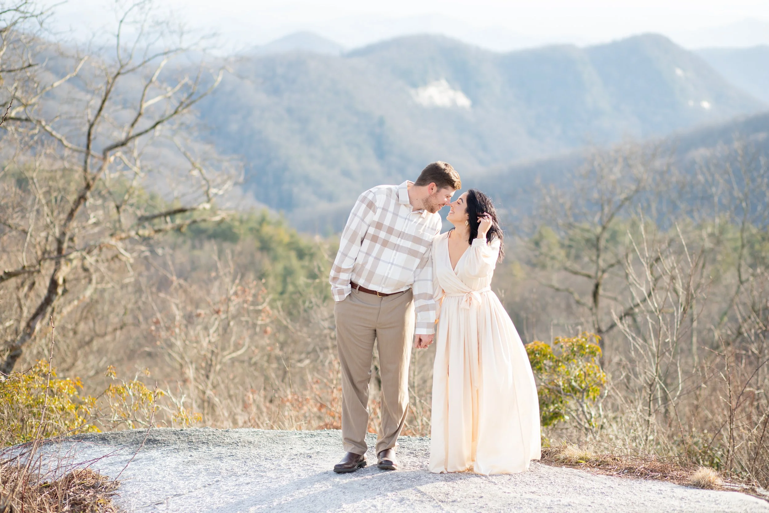 A man and woman holding hands outdoors, standing on a rocky surface, with mountains and leafless trees in the background, during daylight.