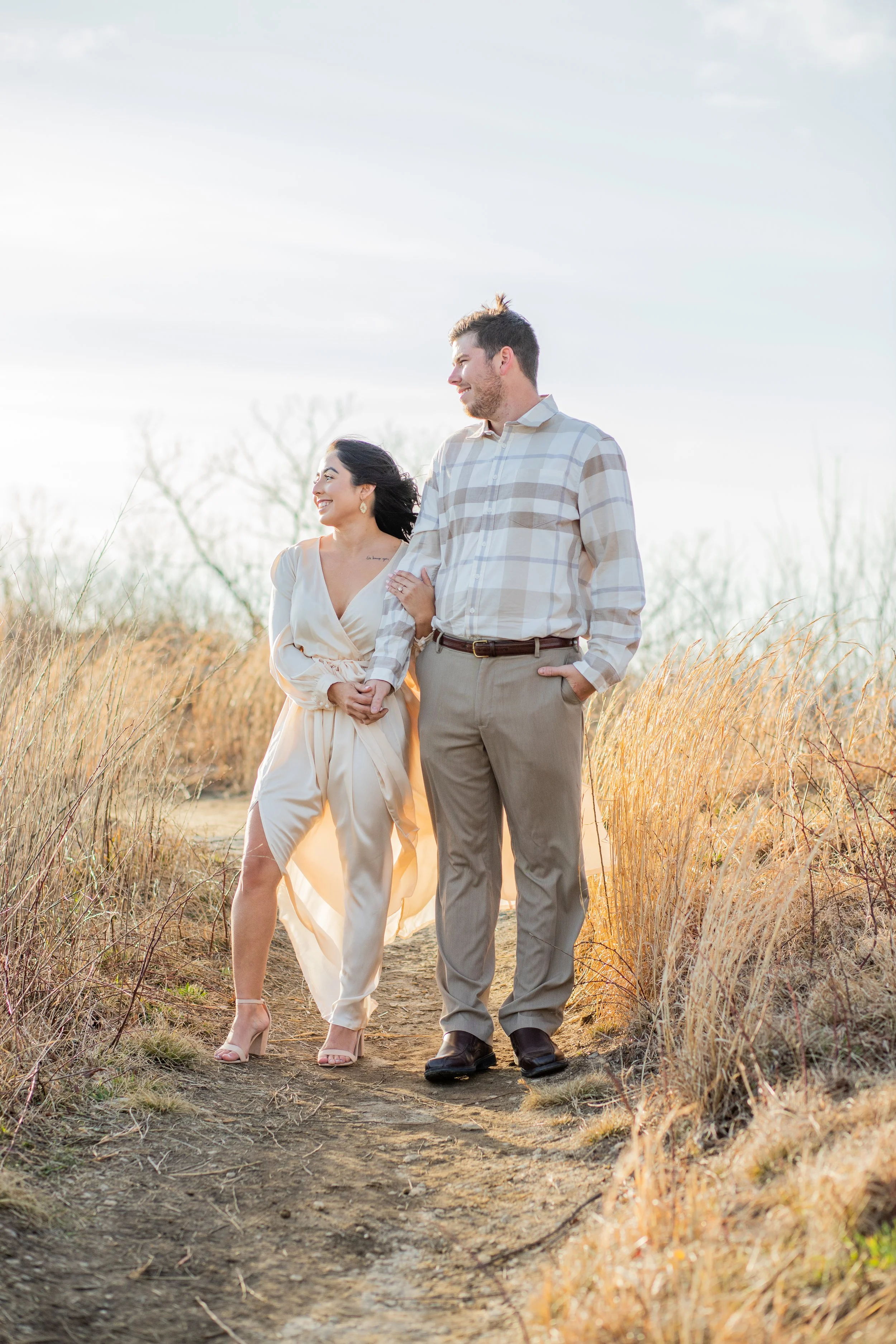A couple walking hand in hand on a dirt path through a grassy field during sunset, smiling at each other.