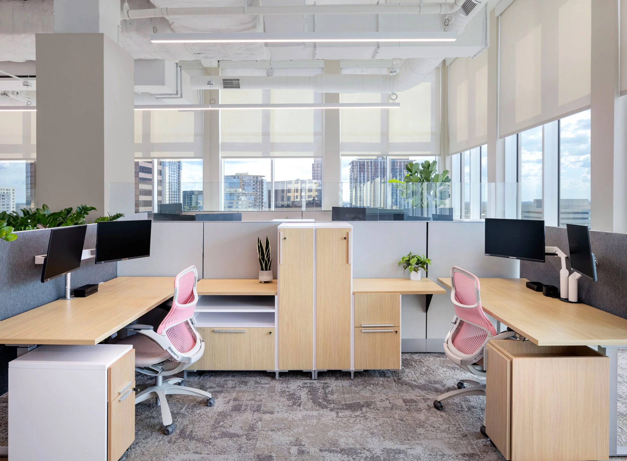 Modern office space with multiple desks, ergonomic chairs, large windows, plants, and city skyline view.