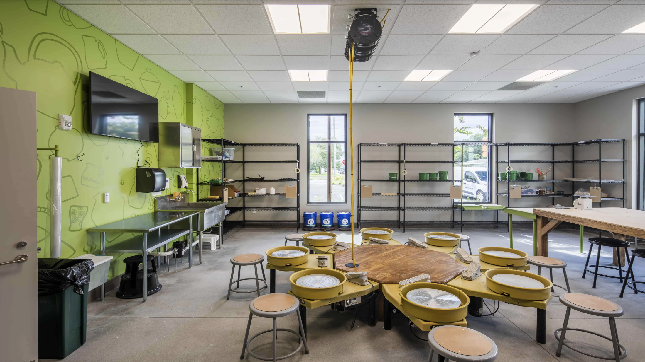 Empty retail space with metal shelving units, a green accent wall, and yellow pottery wheels arranged around a wooden table, indicating pottery or ceramics studio.