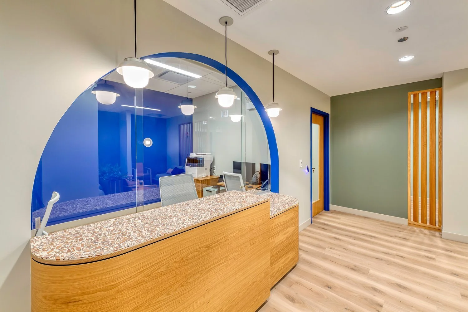 Office reception area with a wooden curved counter, terrazzo top, and a glass wall with a blue semi-circle design. Inside the room are computer monitors, chairs, and office equipment. The space features hanging pendant lights, a green accent wall, and a door with a blue frame.