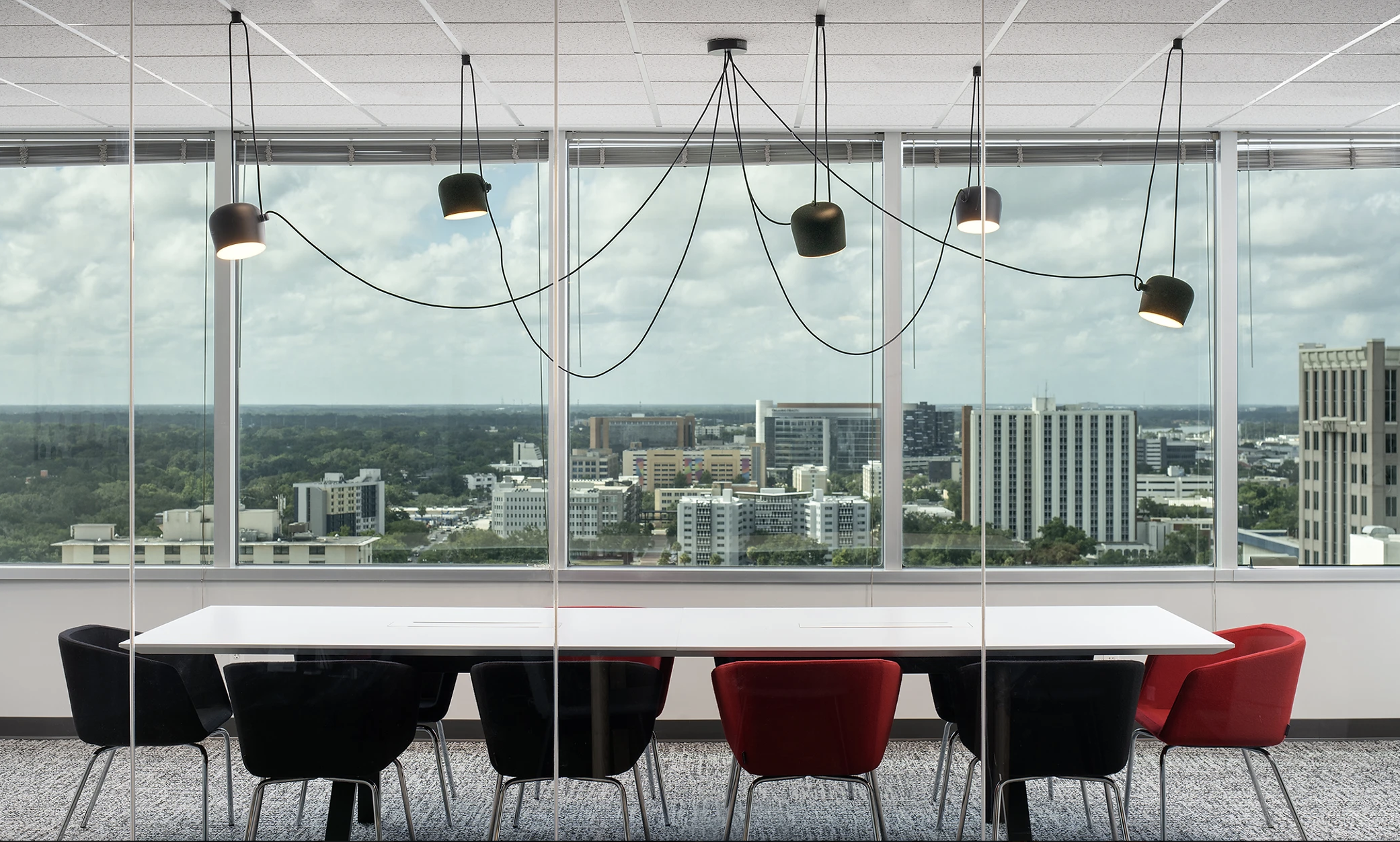 Modern office conference room with large window showing cityscape and sky, black and red chairs around white table, hanging black lamps with cords.