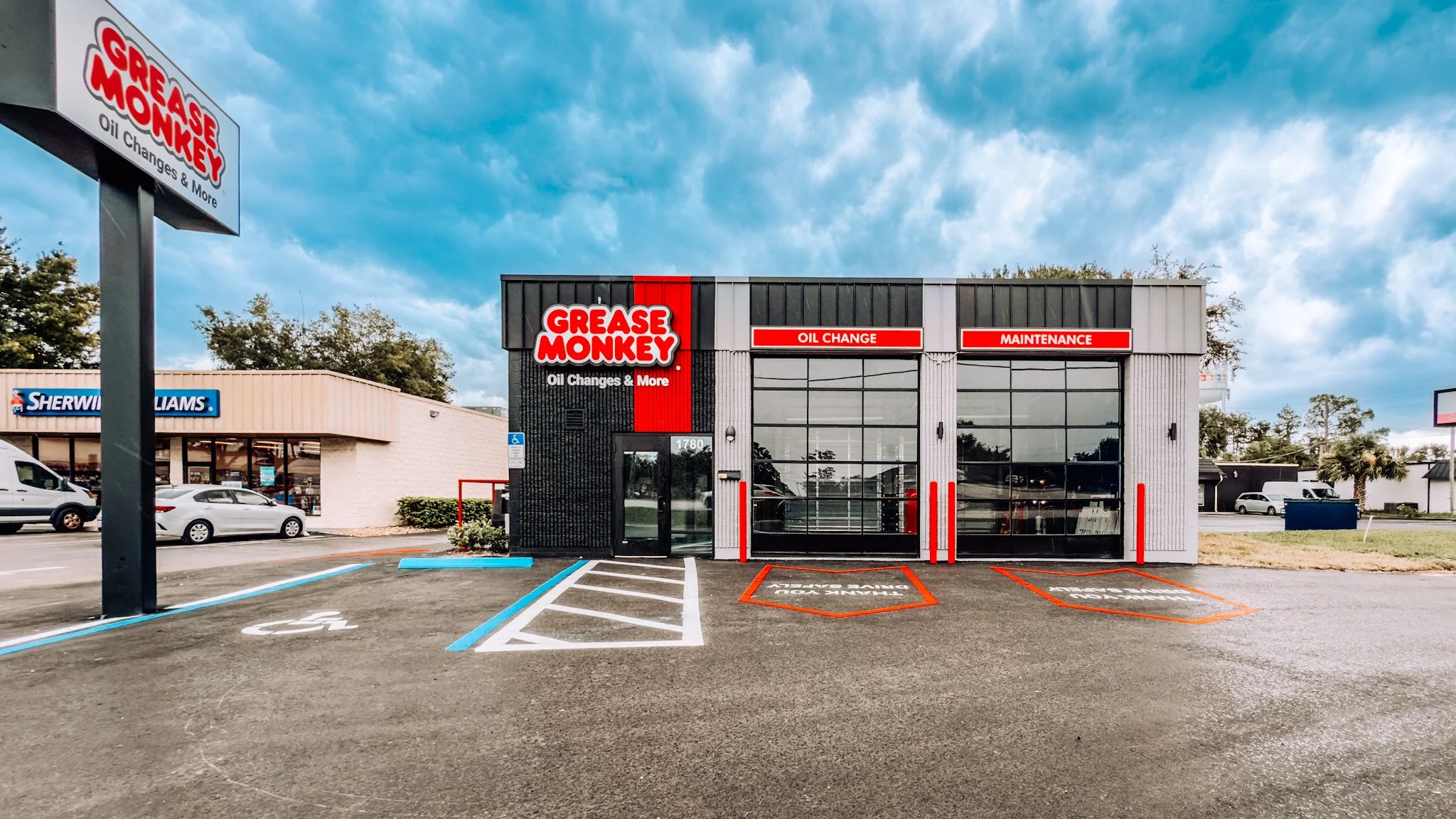 Front view of a Grease Monkey auto service shop with parking spaces in front, including a designated handicapped spot, and a neighboring Sherwin Williams store. The Grease Monkey building has signage indicating oil changes and maintenance services.