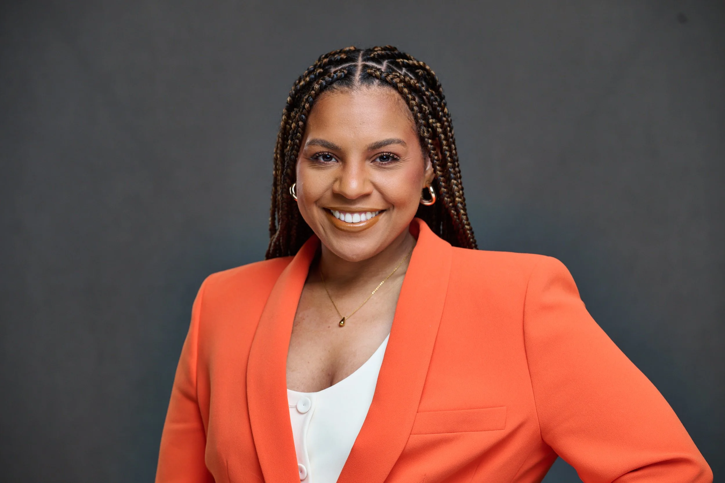 Woman with braided hair smiling, wearing an orange blazer and white top, standing against a dark gray background.