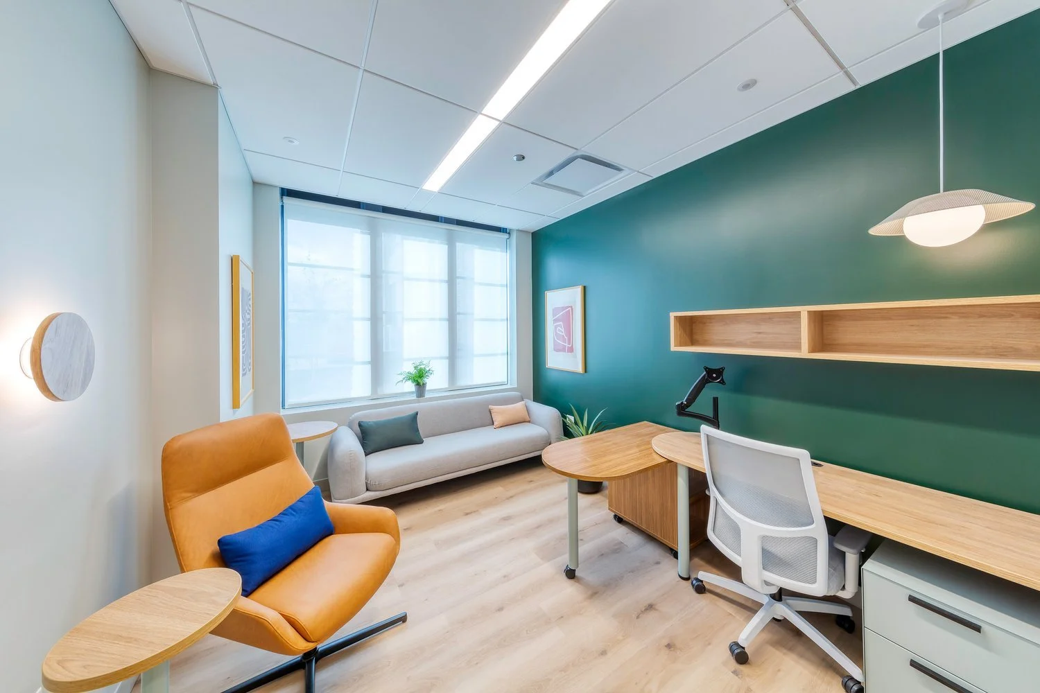 Modern office with a green accent wall, white sofa, orange chair with blue pillow, desk with white office chair, bookshelf, framed art, window with blinds, and plants.