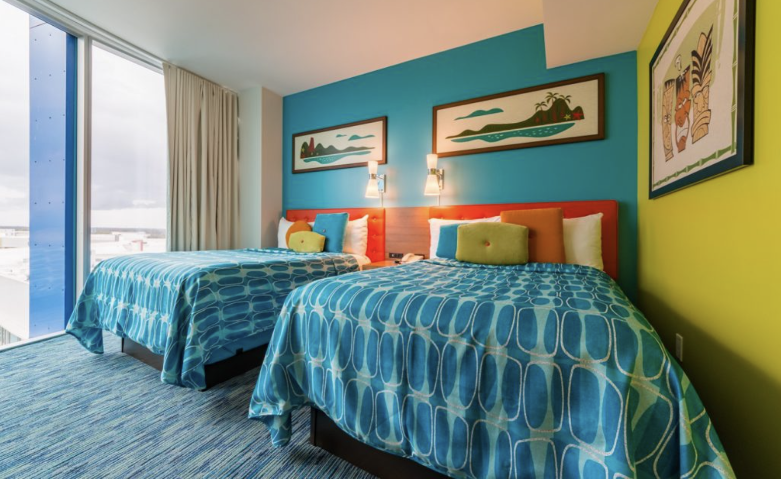Colorful hotel room with two single beds, blue and green bedsheets, decorative pillows, wall art, and large window with curtains.