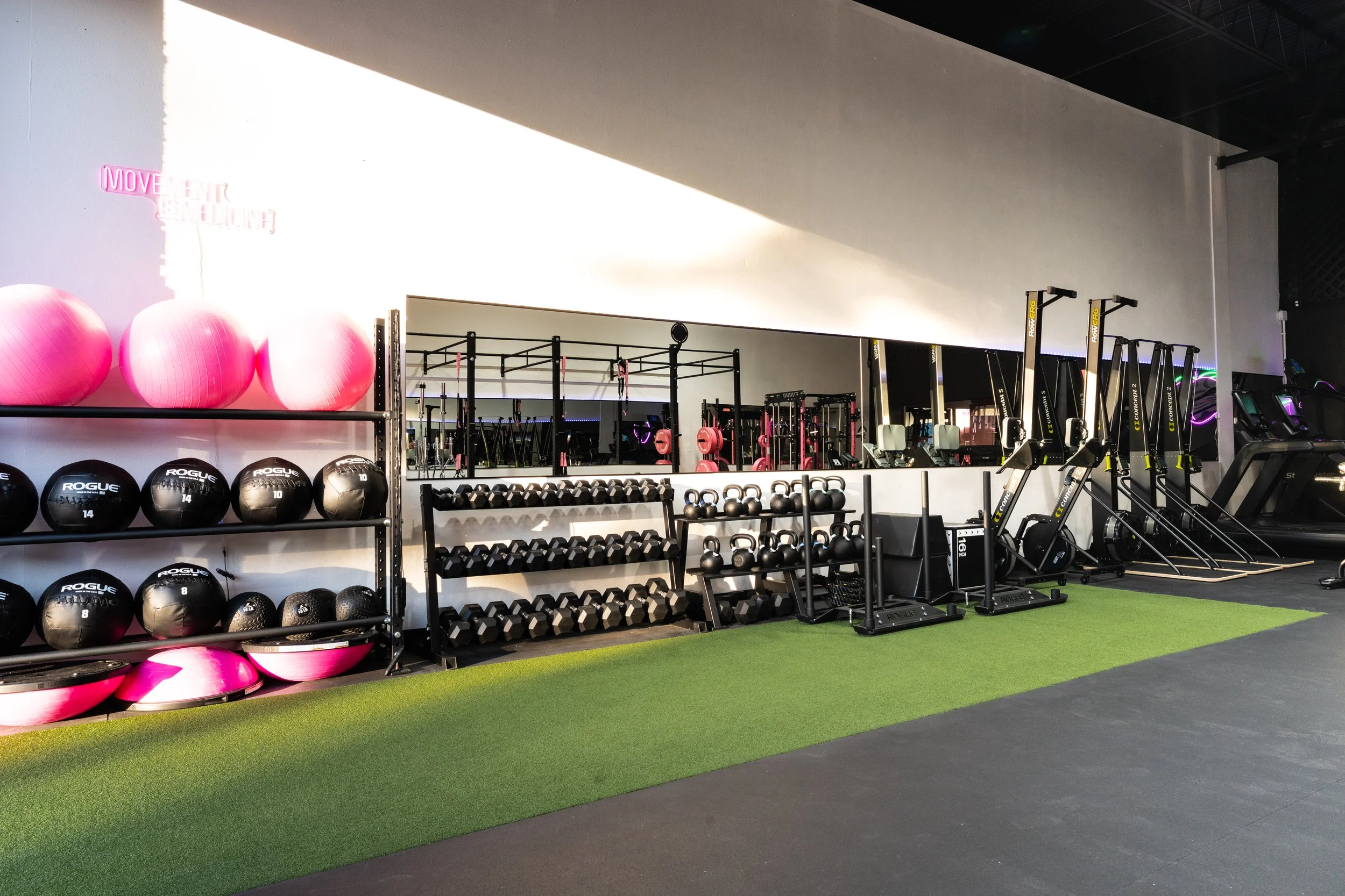 An indoor gym with pink exercise balls, black medicine balls, various dumbbells, kettlebells, and cardio equipment arranged neatly, a large wall mirror, and a neon sign reading 'Movement is Medicine' in pink.