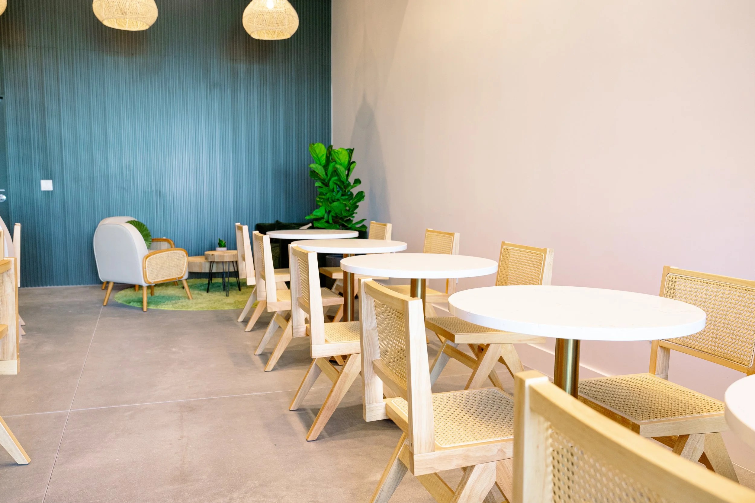 Interior of a modern cafe or lounge area with round white tables, wooden chairs with cane seats, a textured teal wall, a large potted plant, a small side table, and hanging woven lampshades.