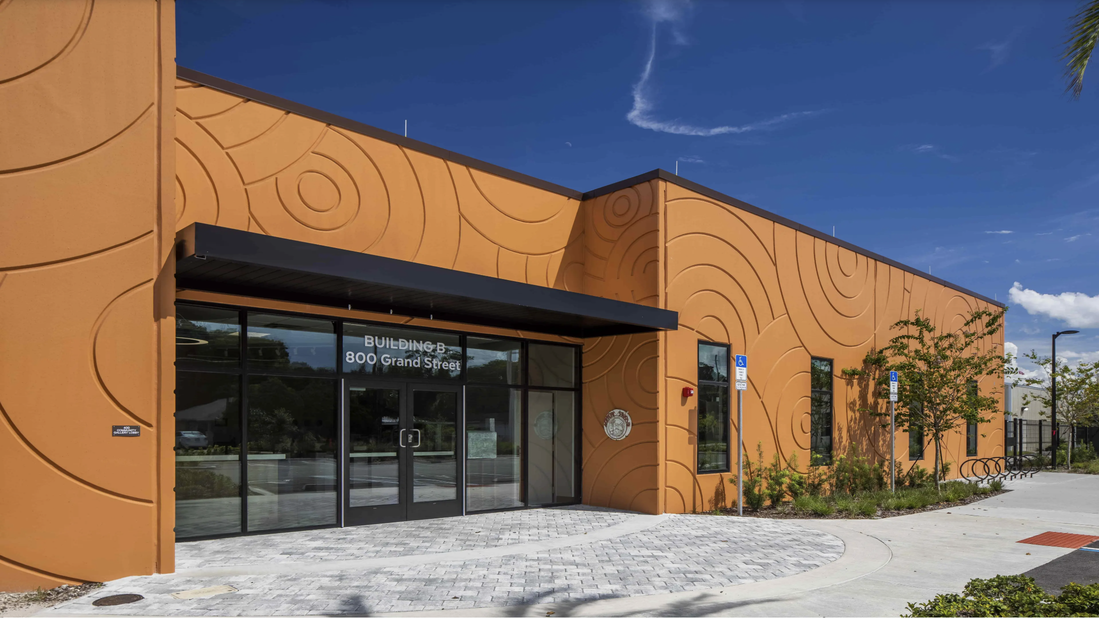 Orange building with decorative circular patterns, glass doors with sign 'Building B 800 Grand Street', surrounding green shrubs, blue sky with some clouds.