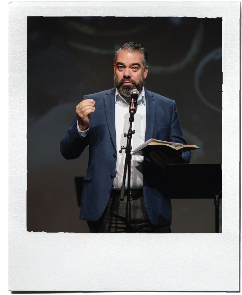 Polaroid-style photo of a pastor speaking into a microphone while holding an open Bible.