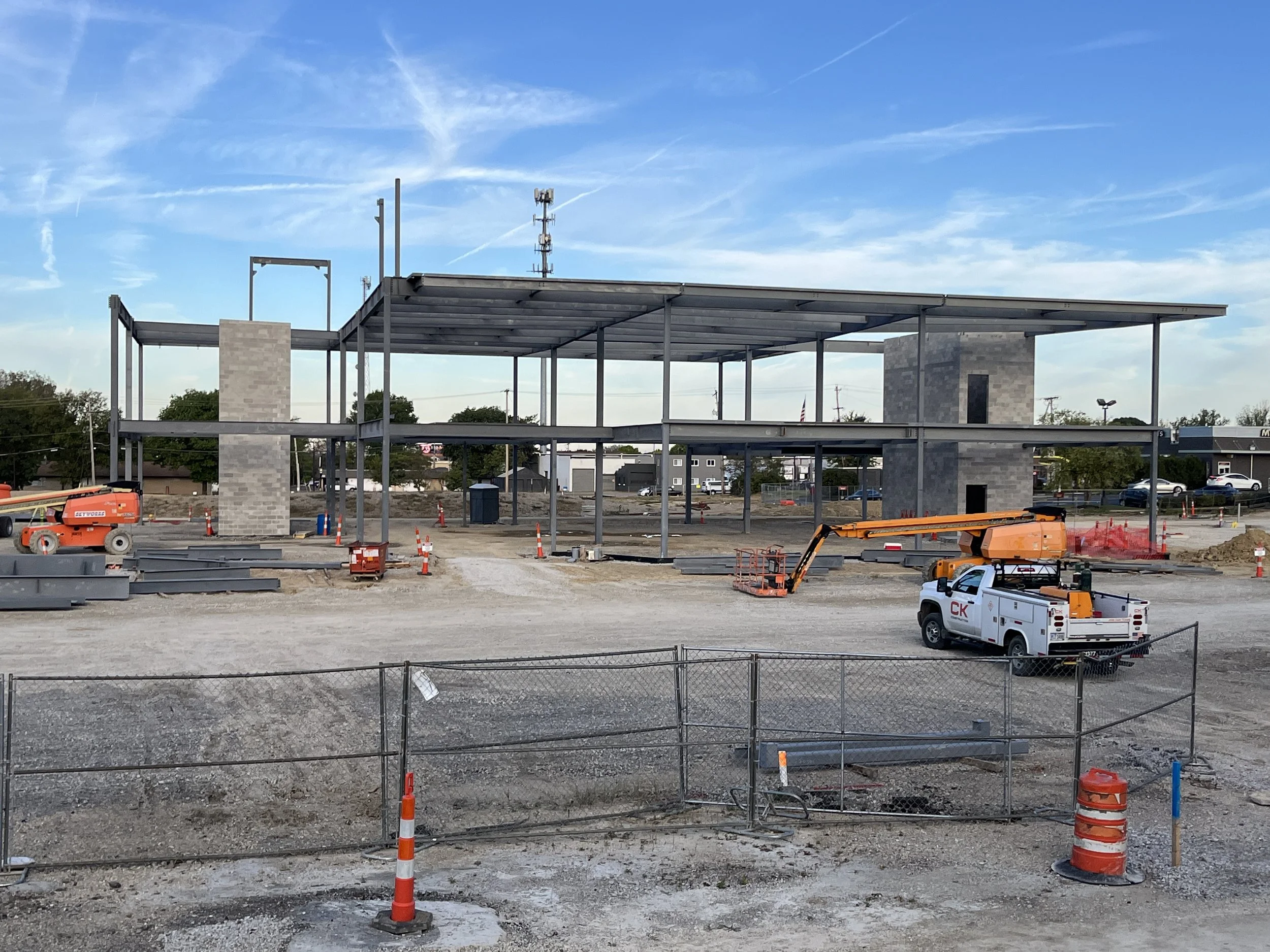 Steel frame of a building under construction with lifts, vehicles, and equipment on a gravel worksite.