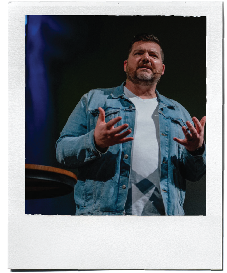 Polaroid-style photo of a pastor speaking on stage beside a podium.
