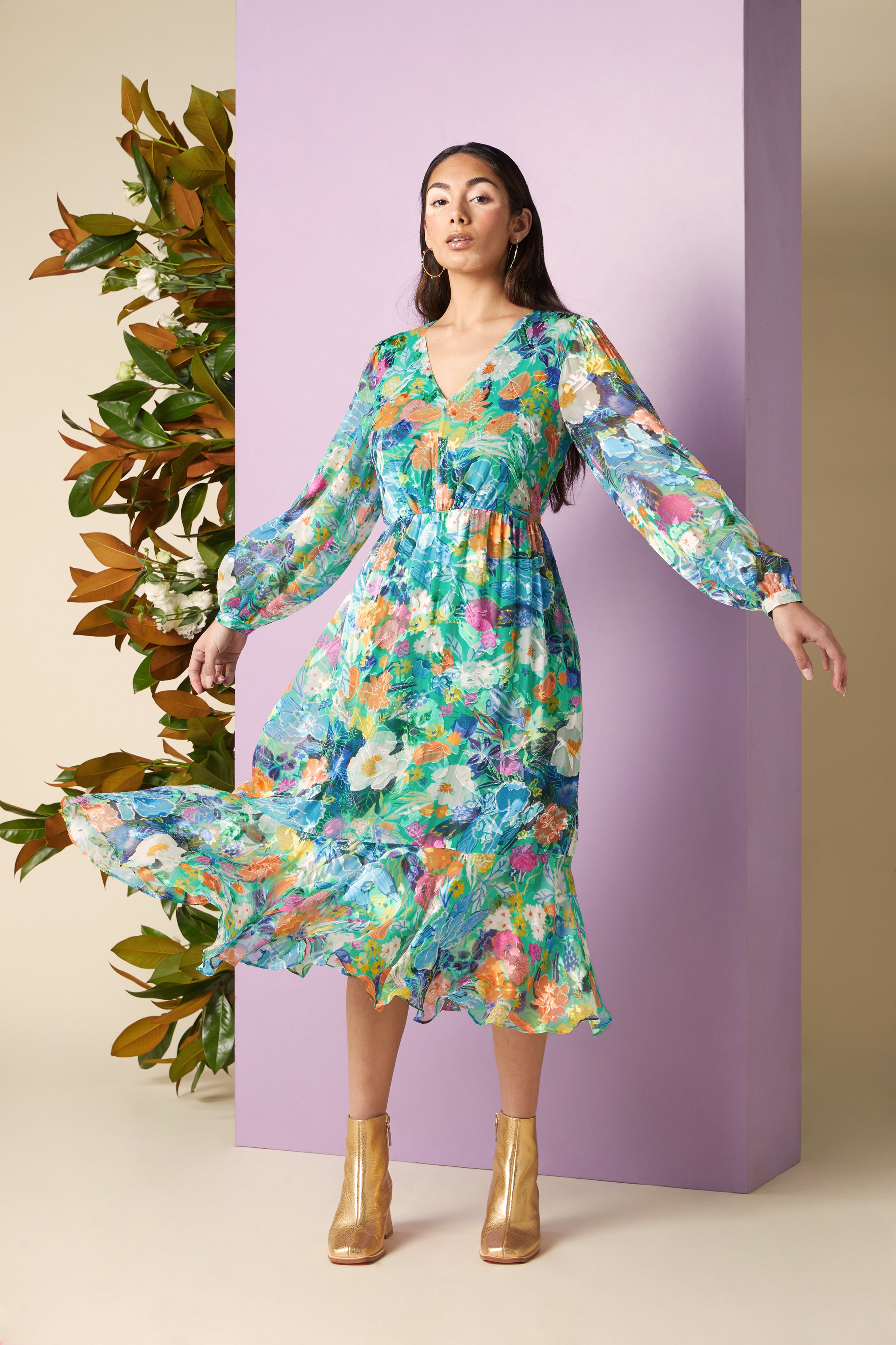 A woman in a colorful floral dress and gold ankle boots poses with one hand extended, standing in front of pastel-colored blocks and a green foliage backdrop.