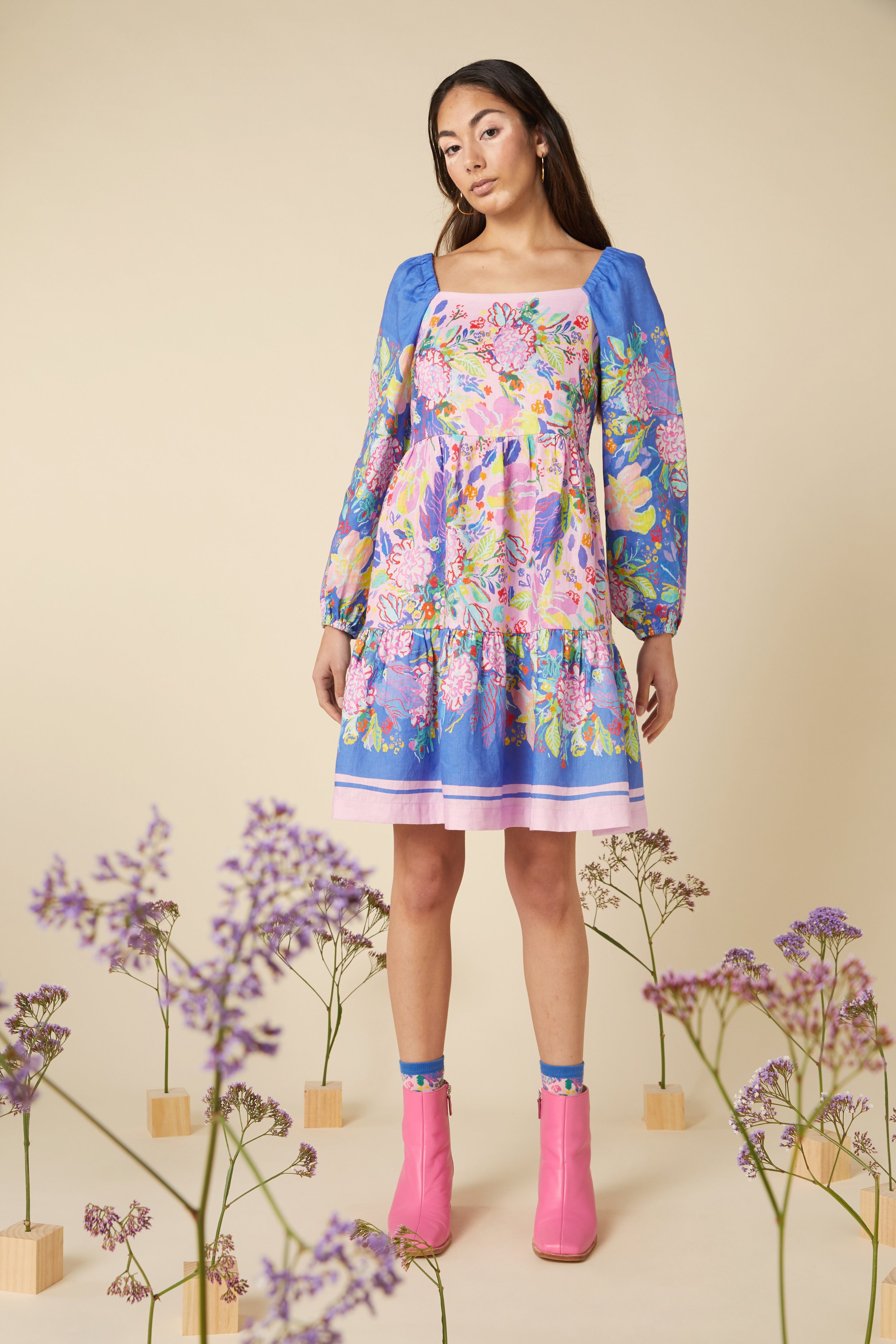 Woman in a colorful floral dress with pink boots standing amidst purple flowers on wooden blocks, against a beige background.