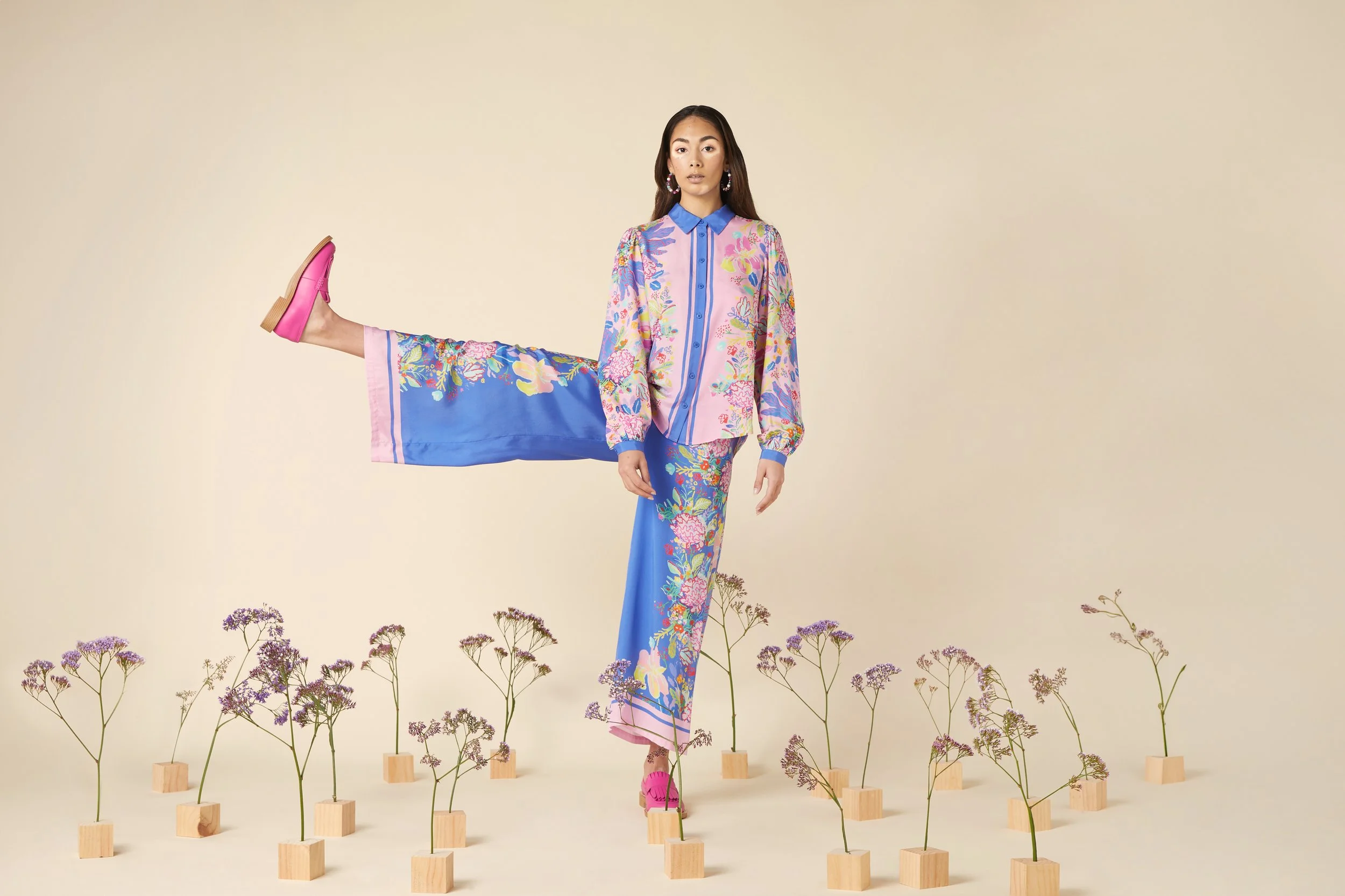 Fashion model standing on one leg, with the other extended to the side, multiple purple flowers in wooden blocks on the ground, wearing a colorful floral outfit, pink shoes, earrings, and a neutral background.