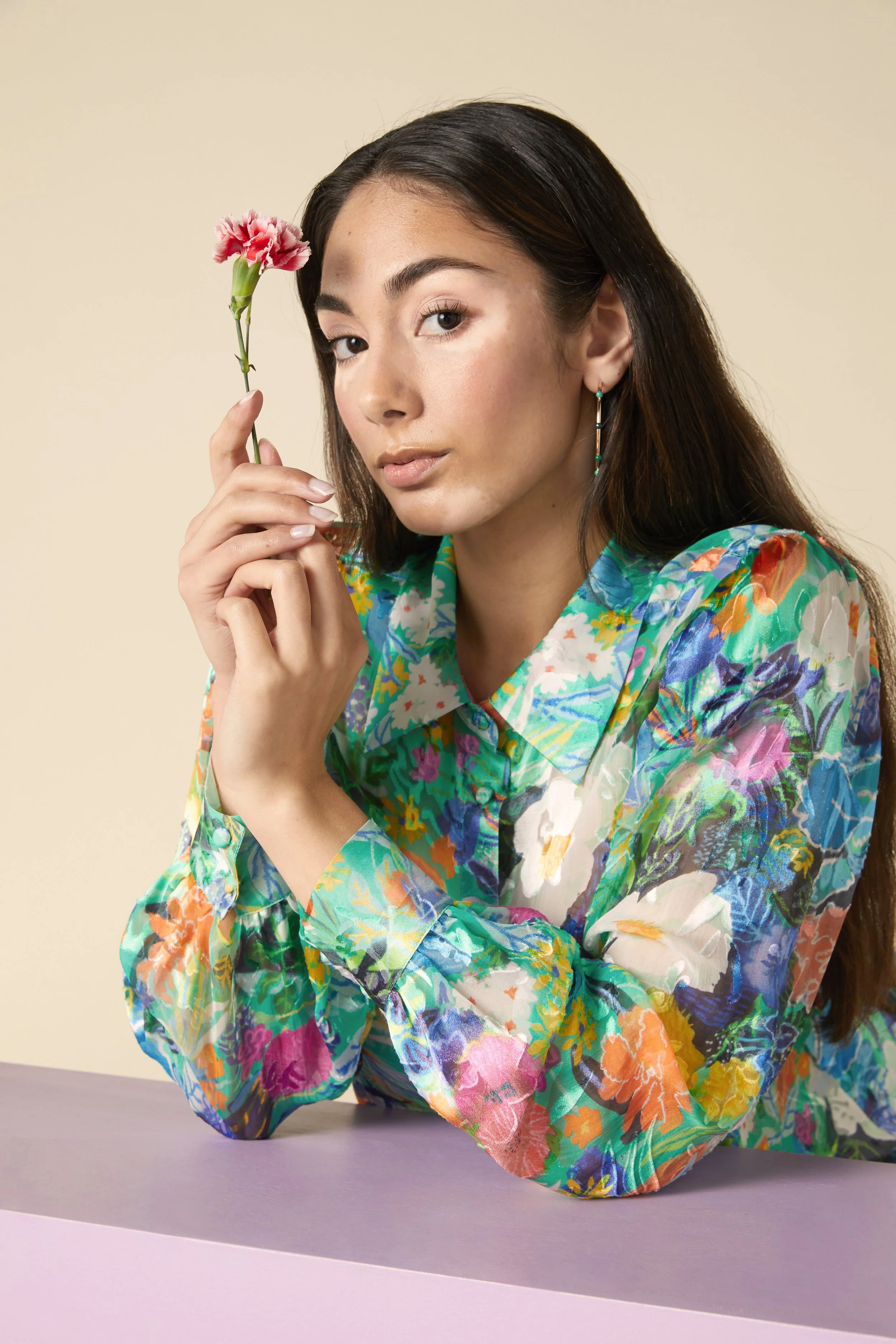 A woman with long dark hair wearing a multicolored floral blouse, holding a pink carnation flower near her face, sitting at a pastel-colored surface against a light beige background.