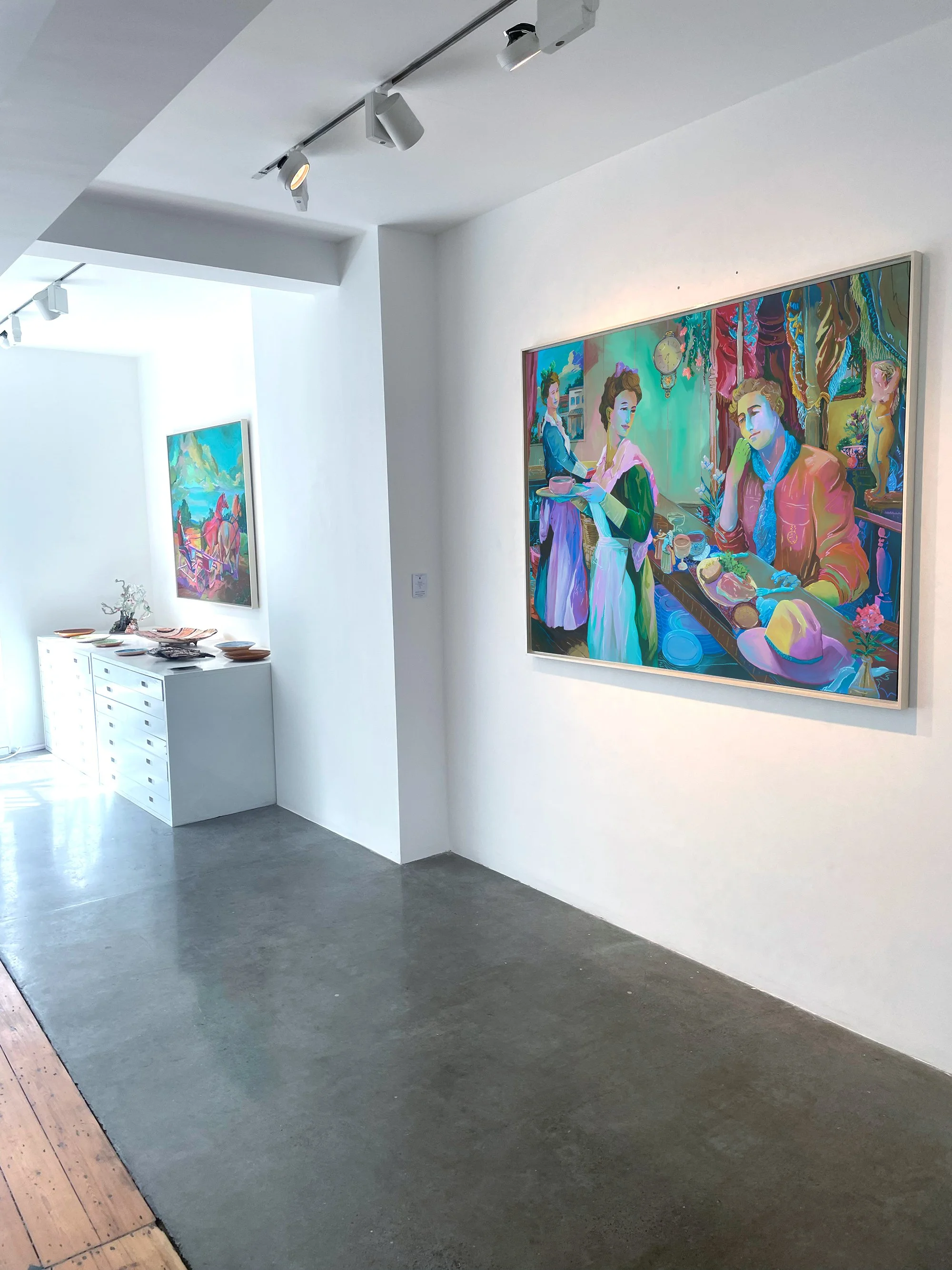 An art gallery with two colorful paintings hanging on white walls. The foreground painting depicts three women in vintage attire in an interior setting. The background painting shows a landscape with mountains, a river, and a bridge. A white console 