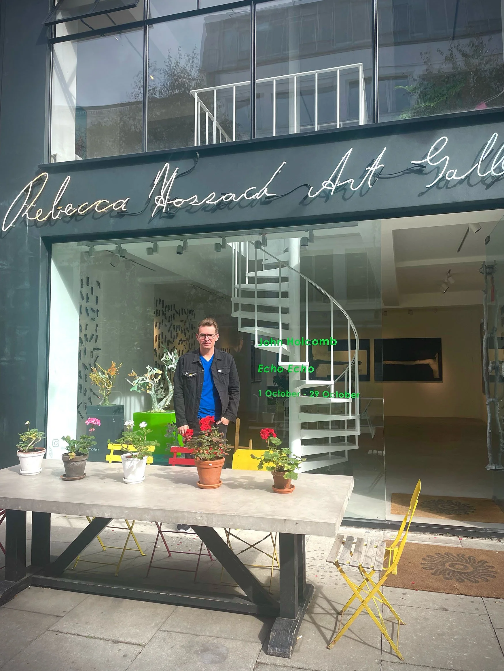 Exterior view of Rebecca Hossack Art Gallery window display, featuring a man standing behind potted flowers on a table in front of the gallery, with a spiral staircase visible inside.