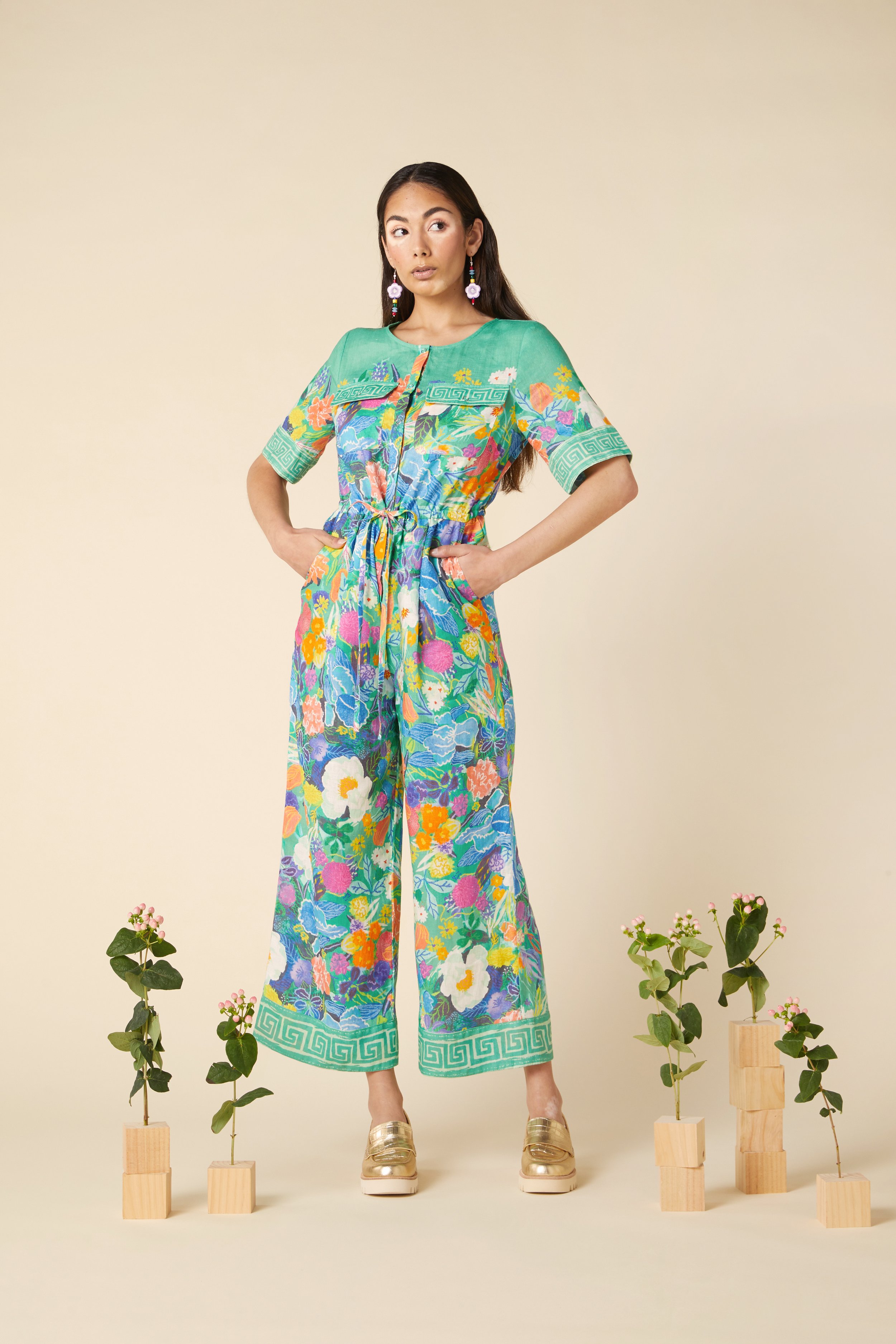 A woman stands with her hands on her hips, dressed in colorful floral and geometric patterned jumpsuit, surrounded by small potted plants with pink flowers.