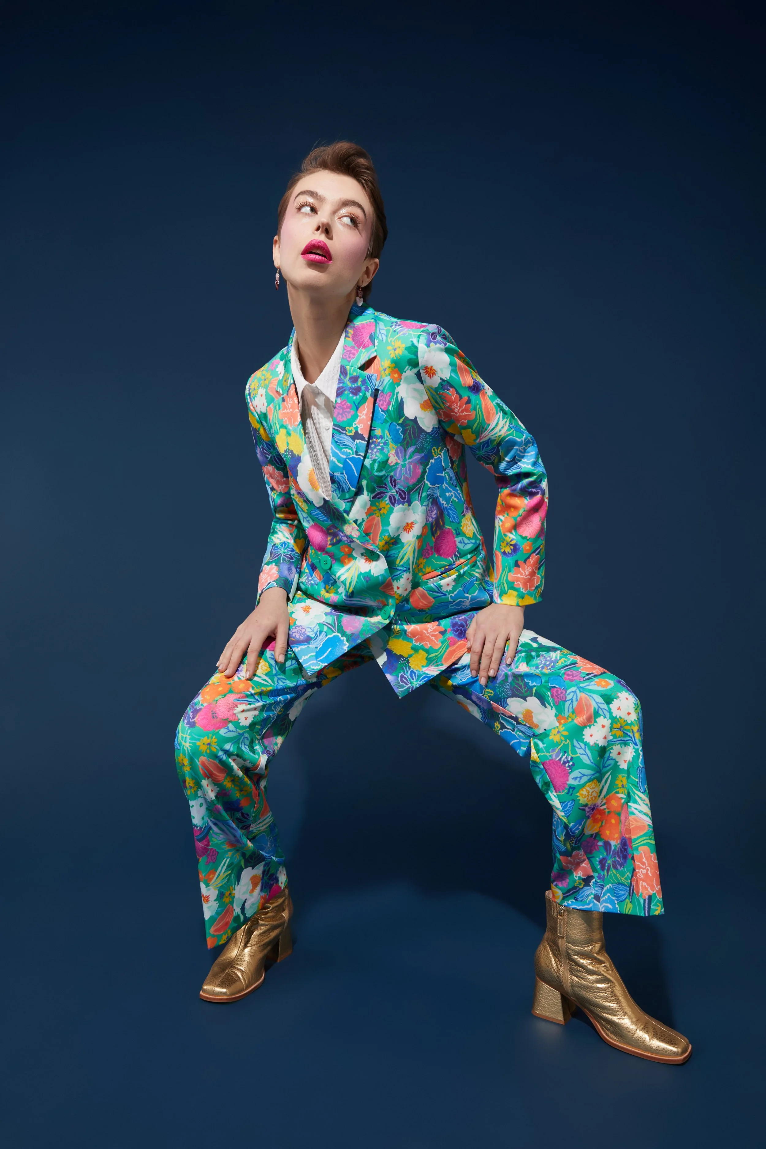 A woman wearing a colorful floral suit and gold boots, posed against a dark blue background.