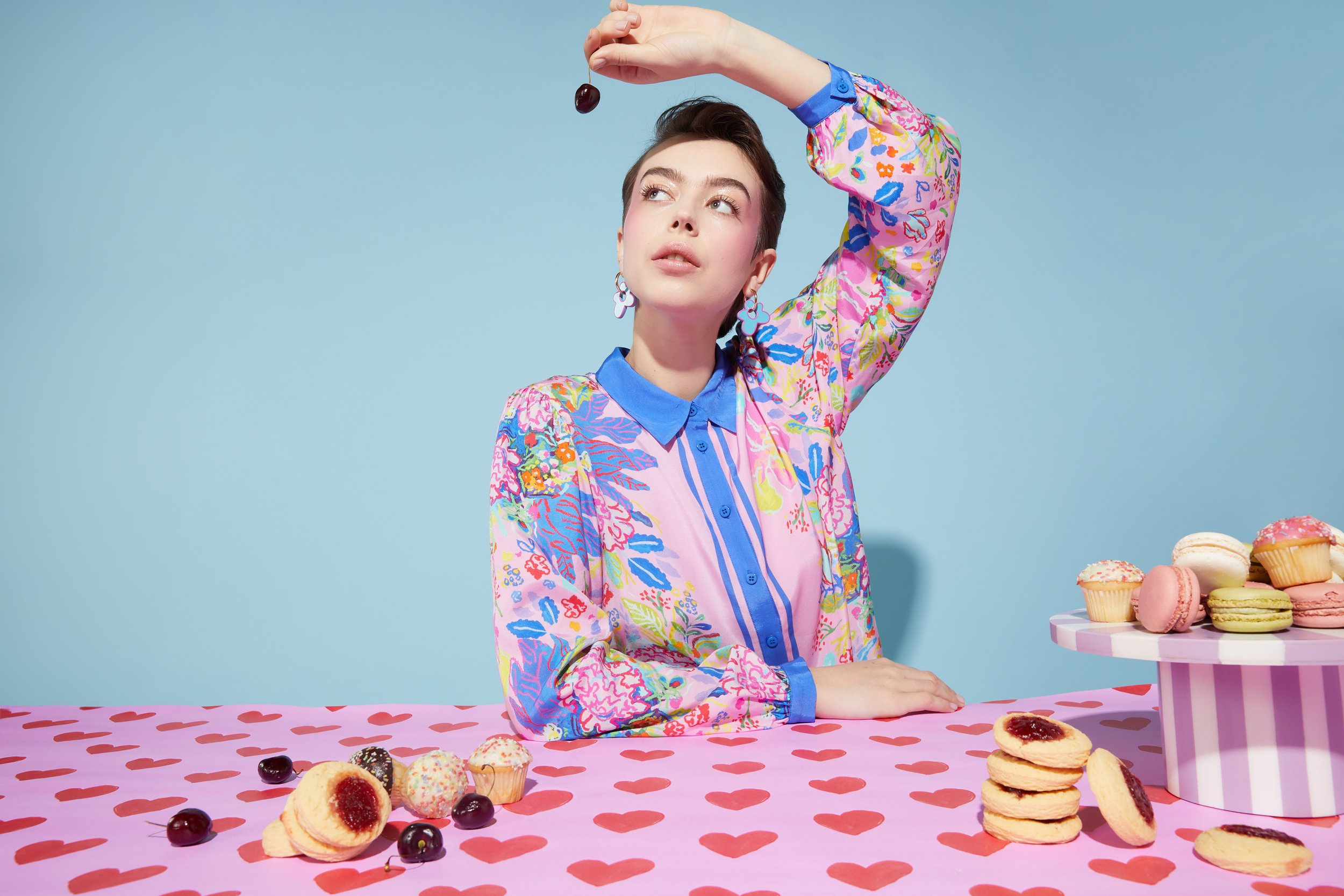 A woman with short dark hair and earrings holding a cherry above her head, sitting at a pink table with a heart pattern, surrounded by cookies, macarons, and cherries, against a blue background.