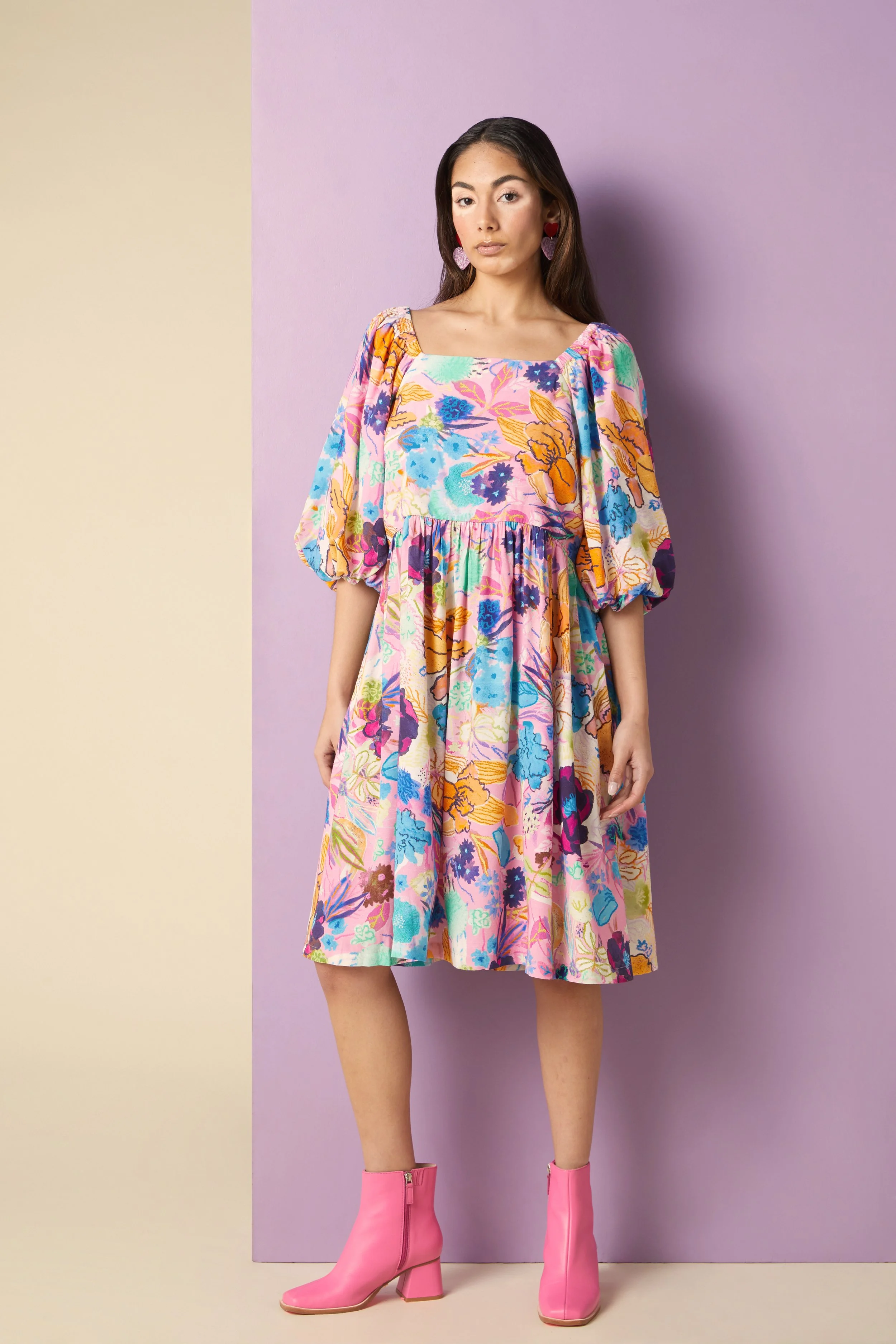 A woman standing against a background with purple and beige sections, wearing a colorful floral dress with puffy sleeves and pink ankle boots.