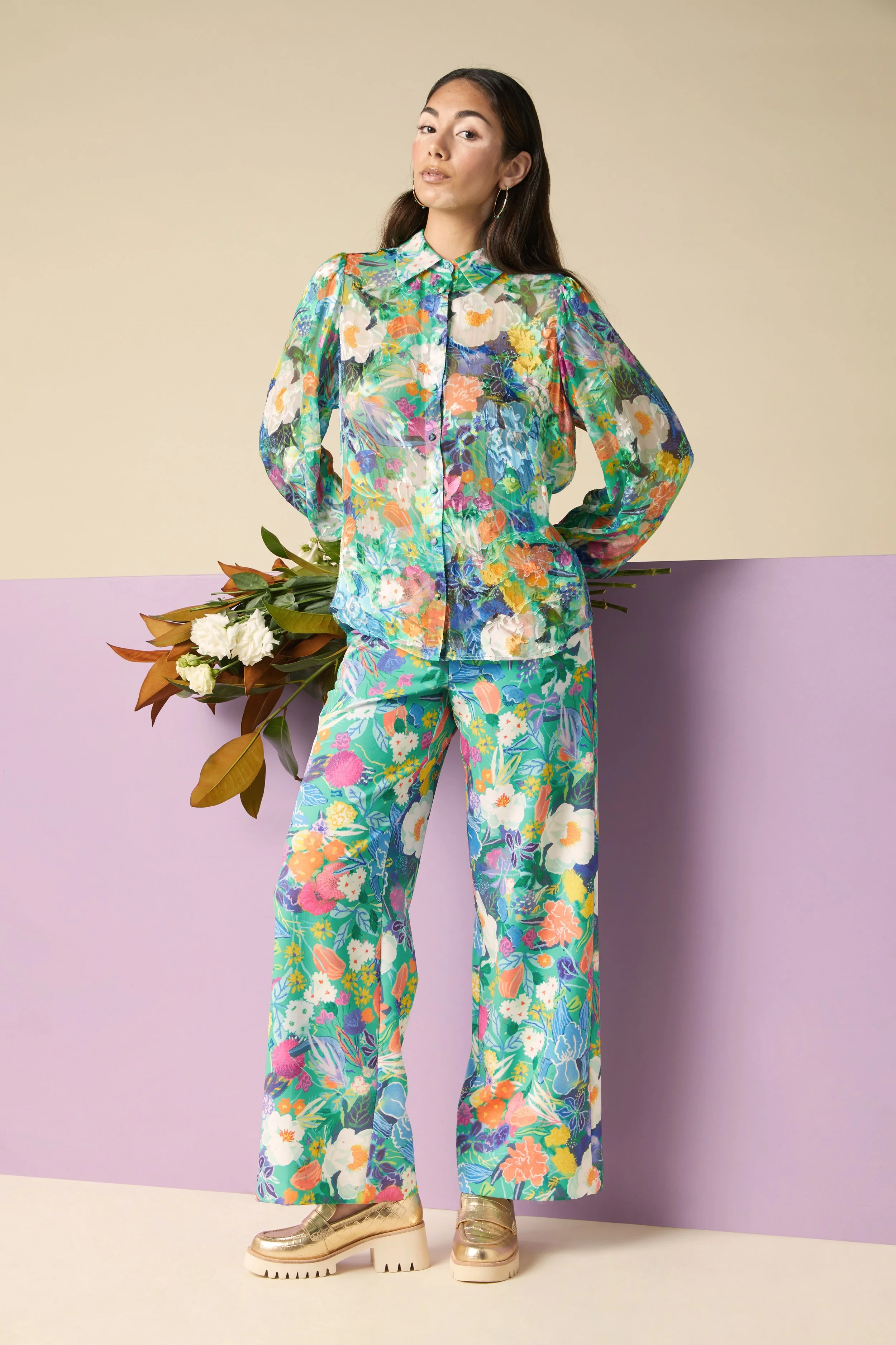 A woman in a vibrant, colorful floral outfit holding a bouquet of flowers, standing against a two-tone background.