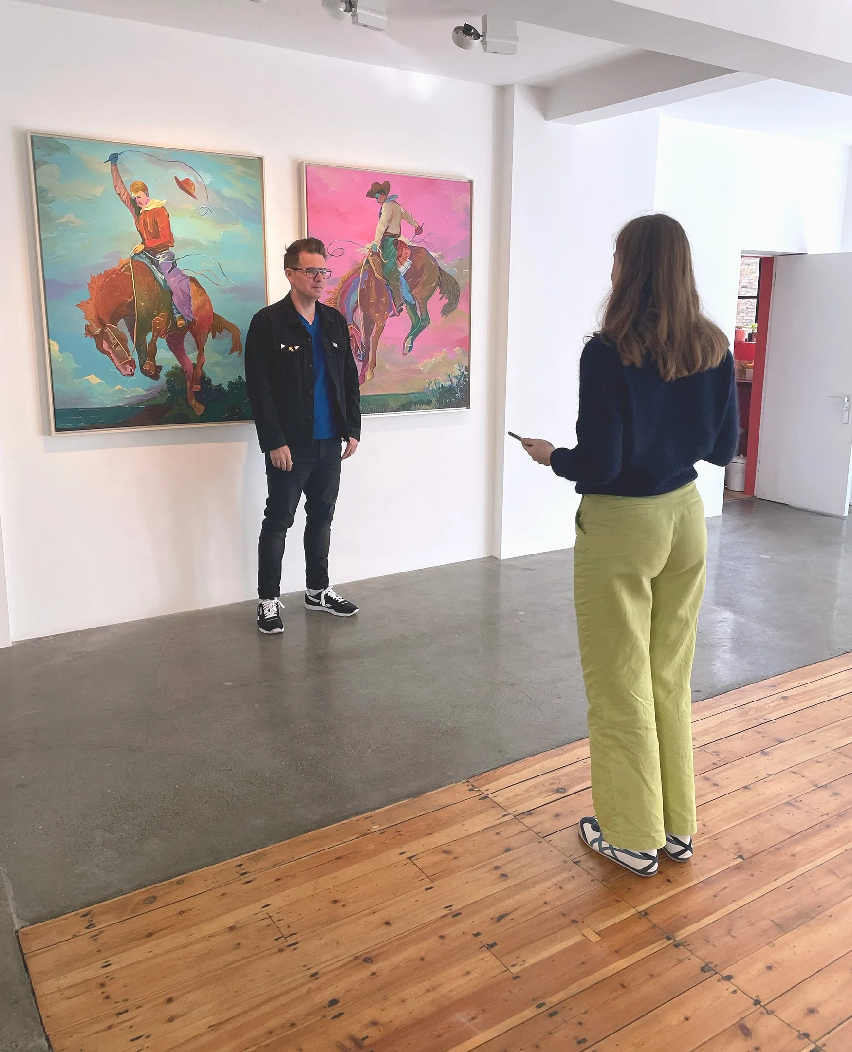 A woman taking a photo of a man standing in front of colorful paintings of cowboys riding horses in an art gallery.