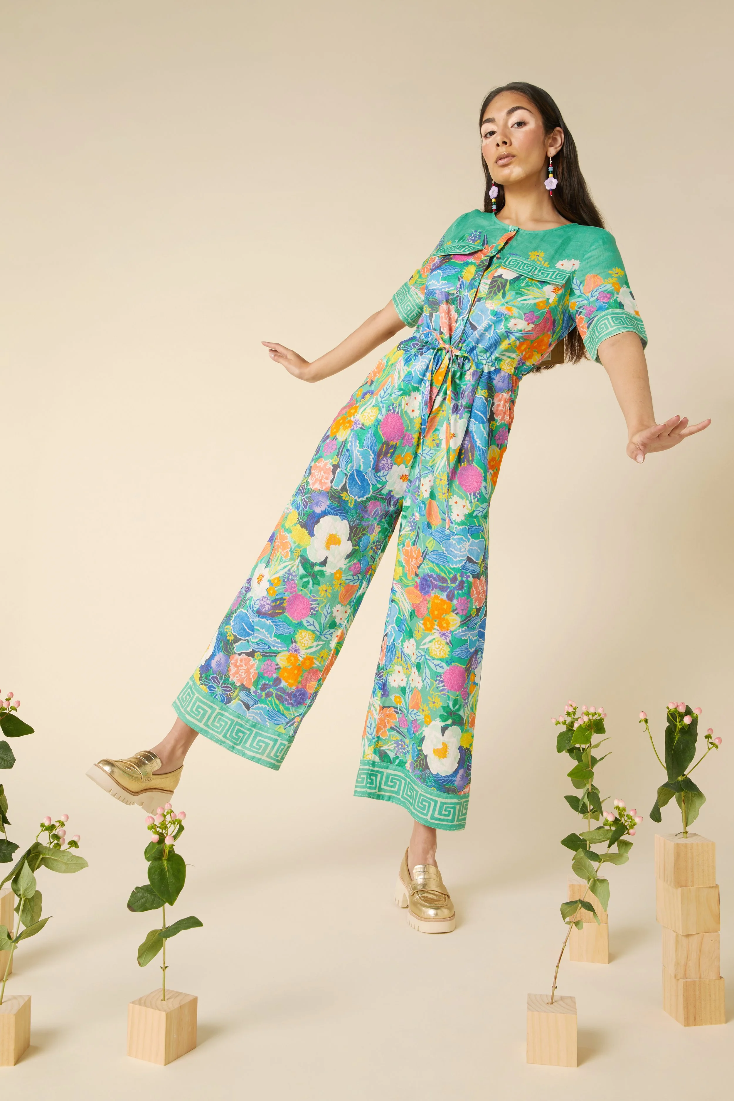 A woman wearing a colorful, floral jumpsuit standing with arms outstretched against a beige background, surrounded by small plants in wooden blocks.