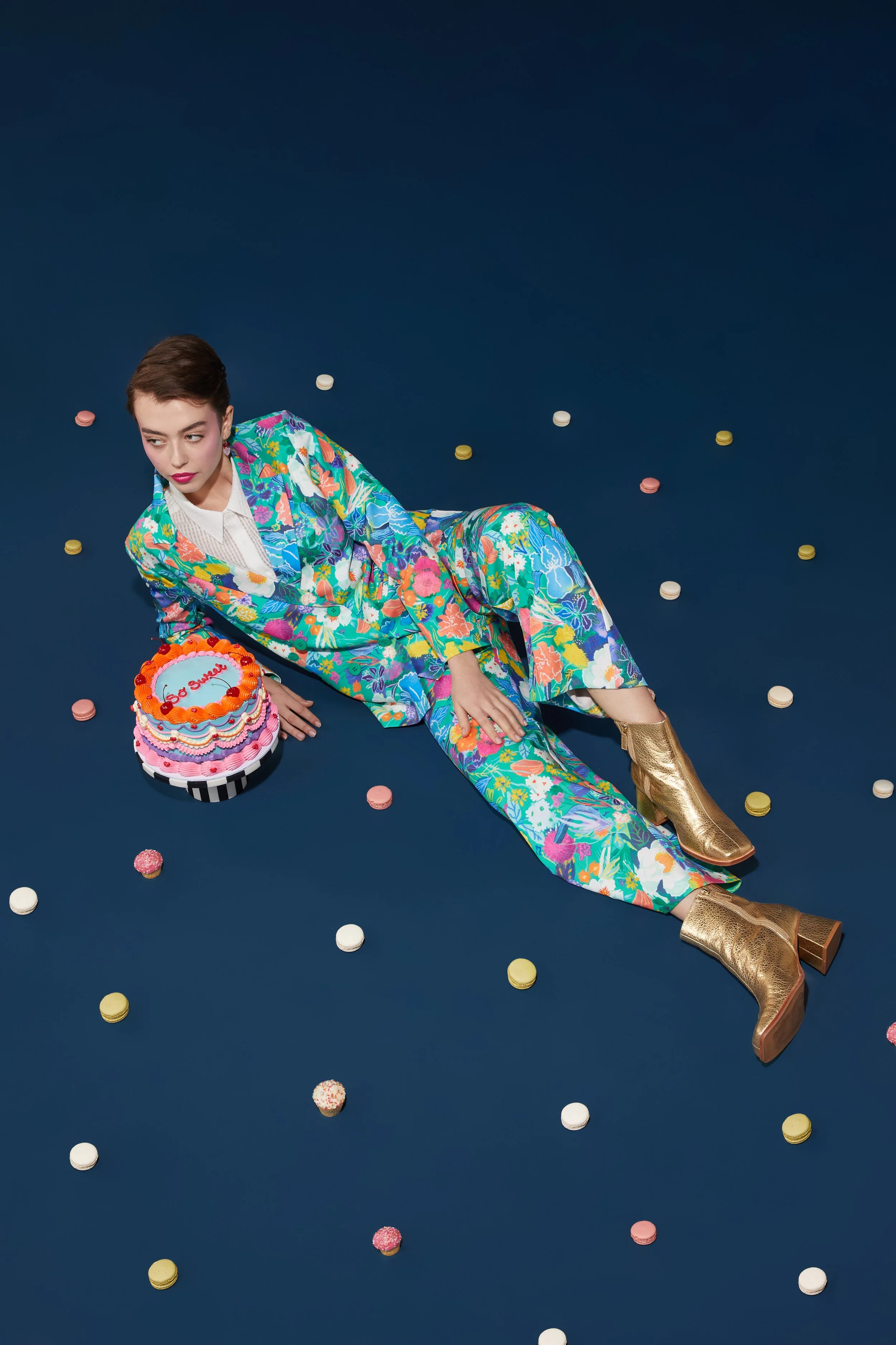 A woman wearing a colorful floral suit and gold boots sitting on the floor surrounded by scattered candies and a decorated cake with a message, on a dark blue background.