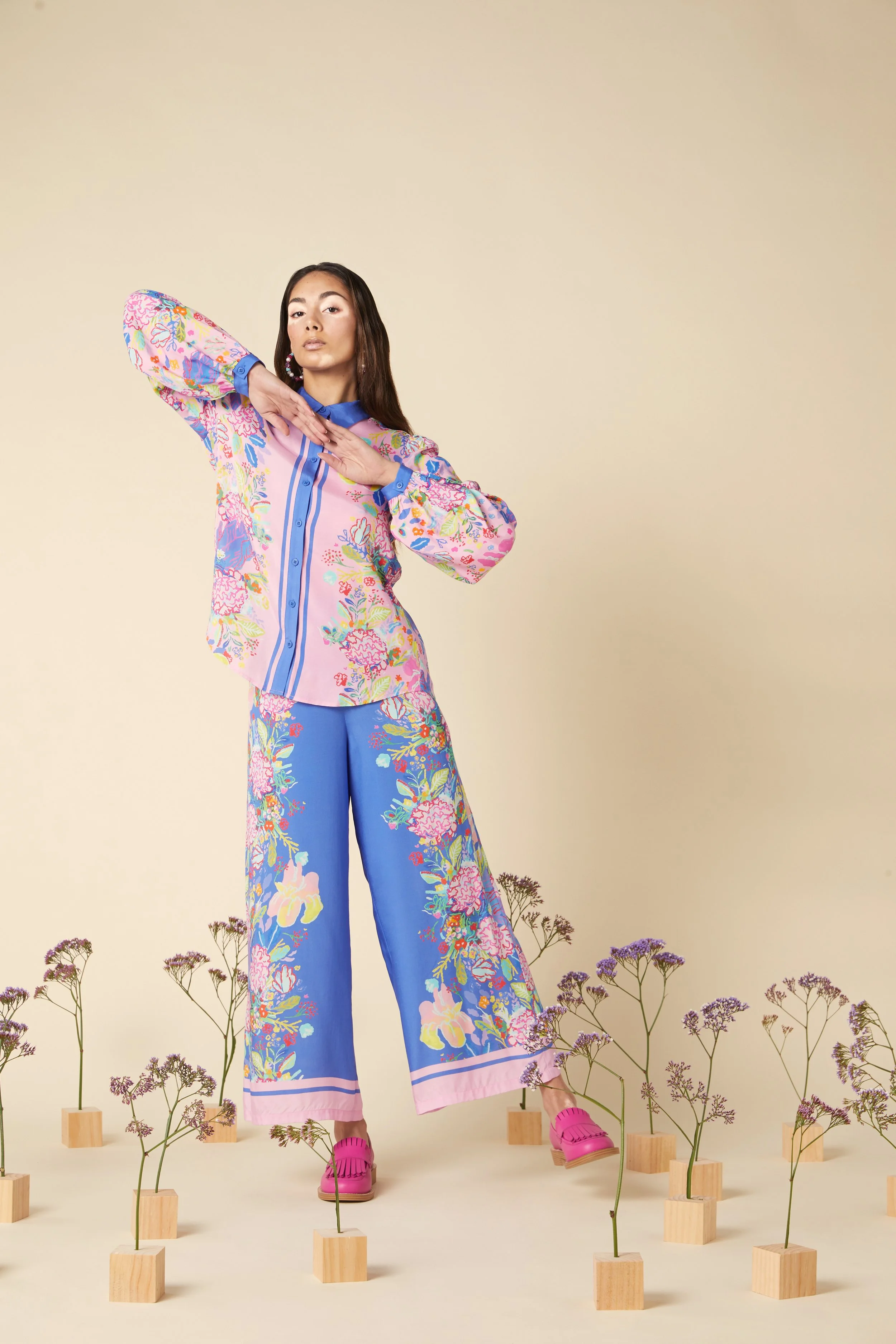 A woman standing among small wooden blocks with purple flowers on top, wearing a colorful floral blouse, wide-leg blue pants with floral pattern, pink shoes, and posing with one arm across her chest and the other bent at the elbow.