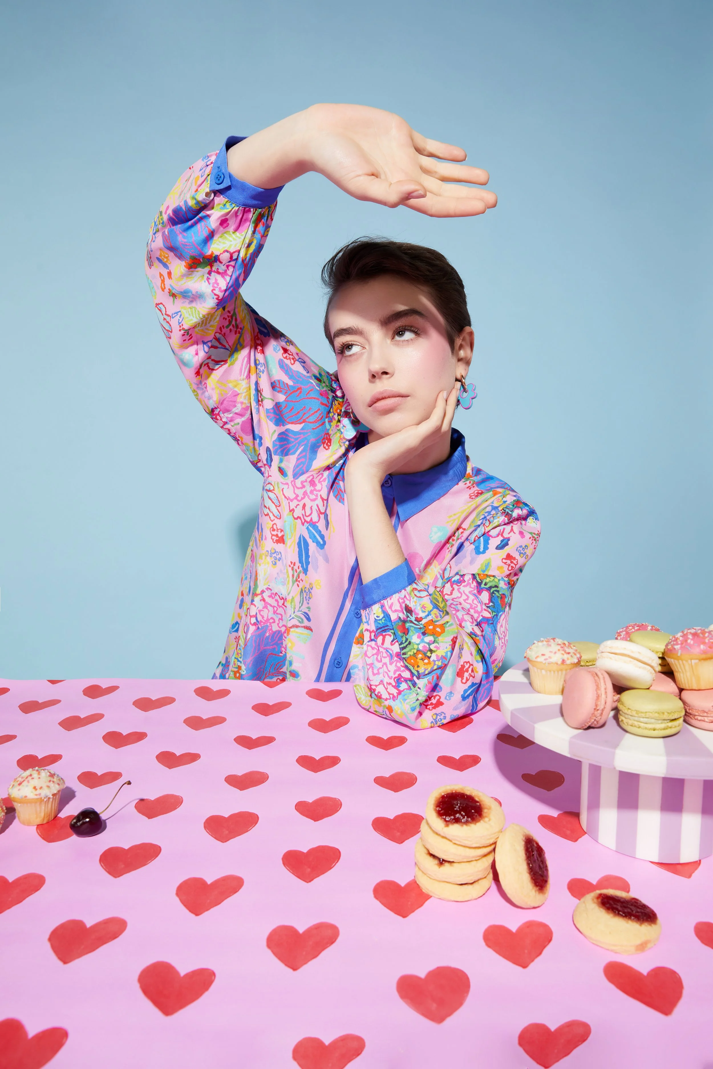 A young person with short dark hair wearing a colorful floral shirt sits at a pink table with red heart patterns. They are resting their chin on their left hand, looking thoughtfully while their right hand is raised above their head. On the table are