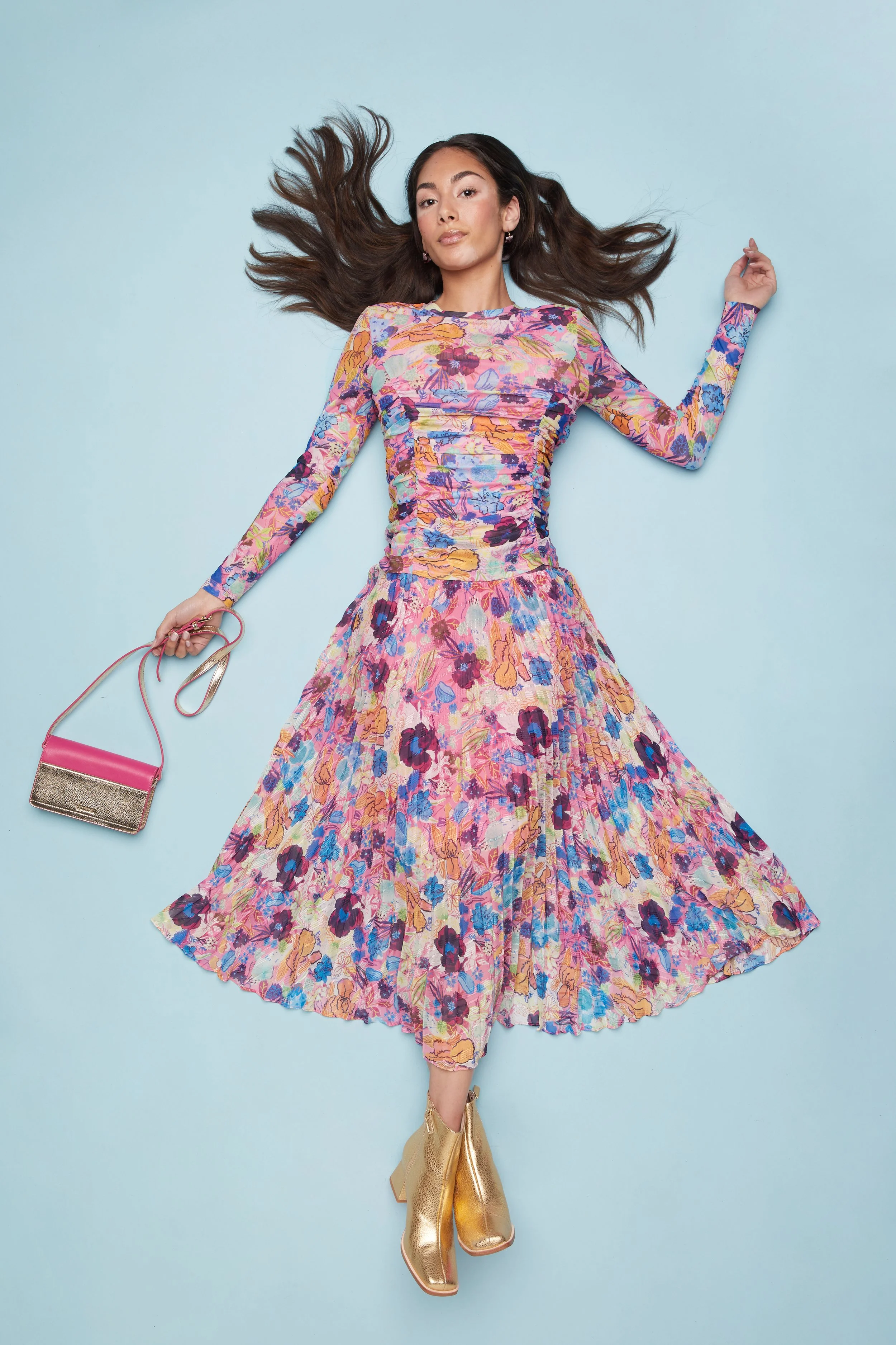 A woman lying on a light blue background, wearing a colorful floral dress, gold ankle boots, and holding a small pink and gold handbag.