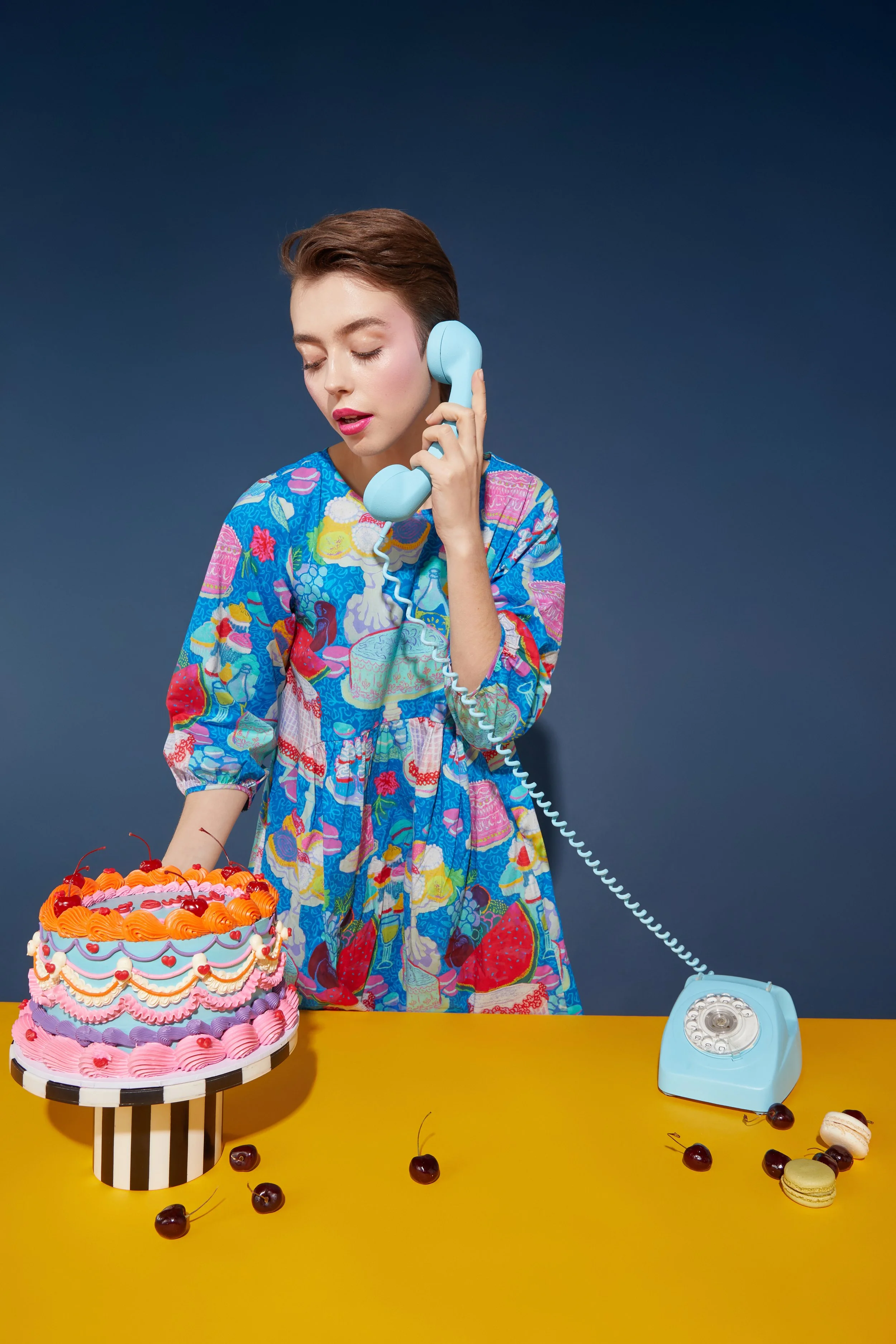 A woman with short brown hair, wearing a colorful dress with fruit and dessert prints, is holding a light blue telephone receiver to her ear. She is standing behind a yellow table with a large, decorated birthday cake, cherries, and macarons, against