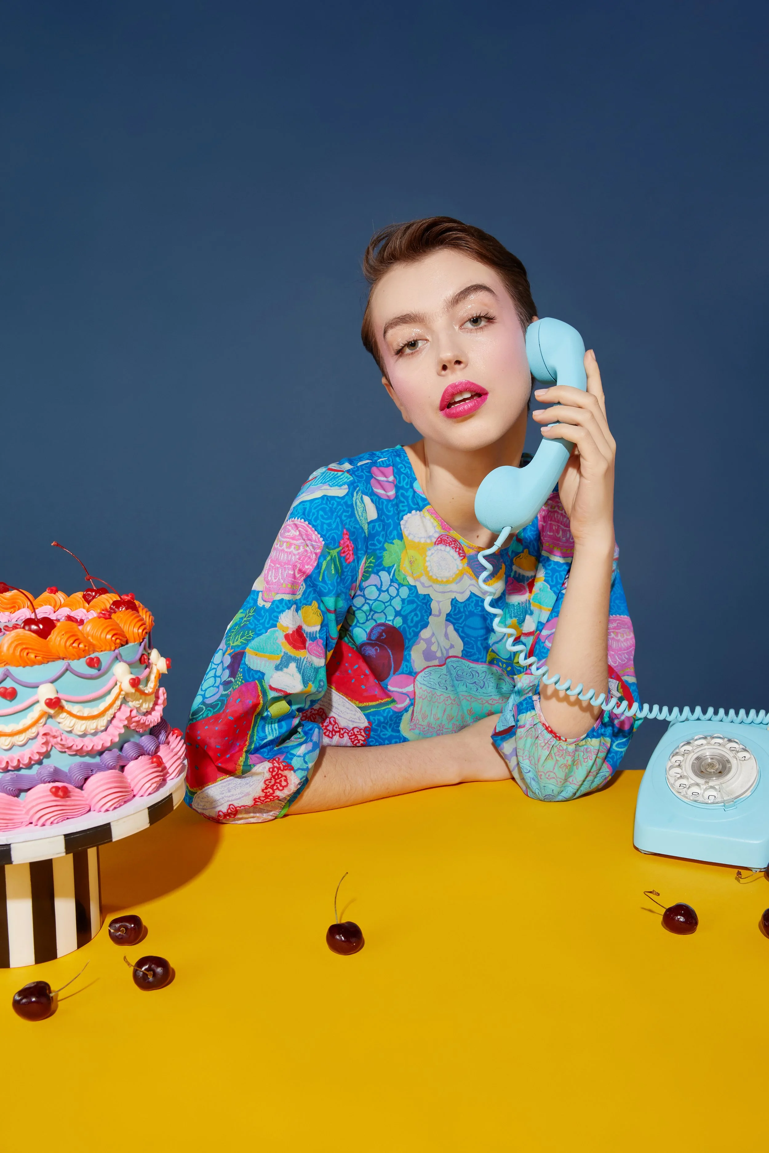 A woman with short brown hair and pink lipstick is holding a light blue rotary phone to her ear, sitting at a yellow table with a colorful cake and cherries. She wears a vibrant, patterned blouse and is against a blue background.