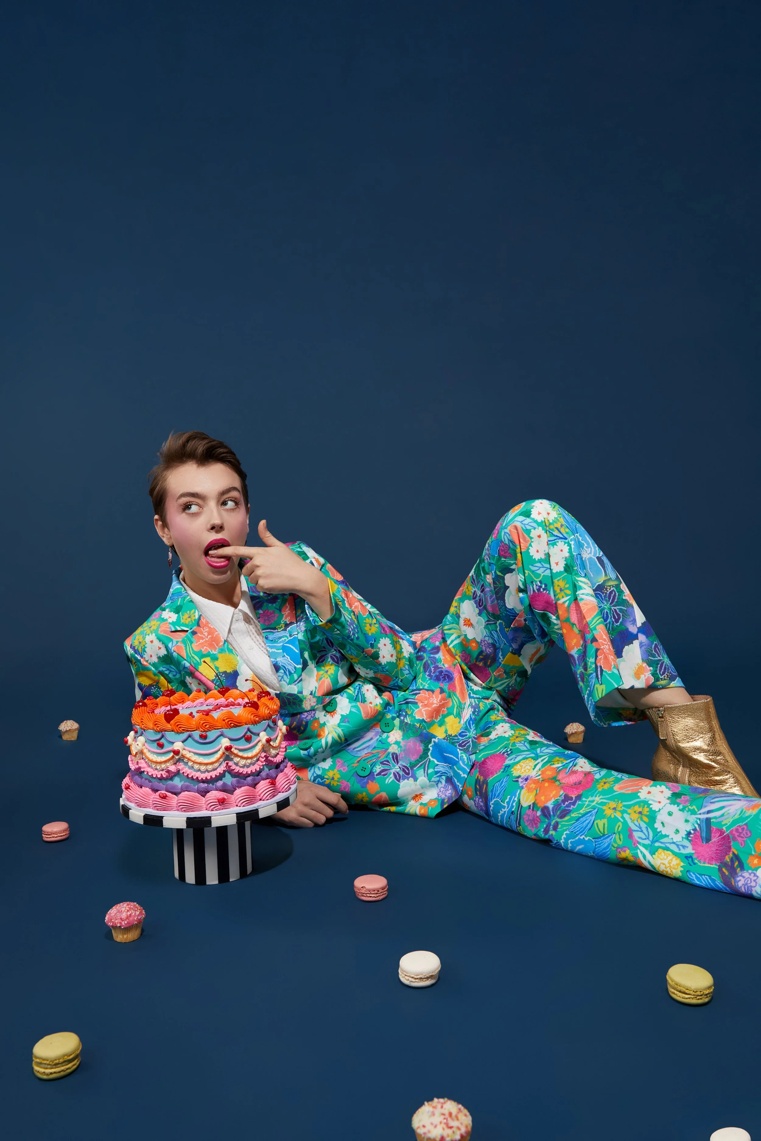A woman in a colorful, floral suit lying on a dark blue surface surrounded by cupcakes and macarons, with a multi-layered pink and orange cake on a striped cake stand.