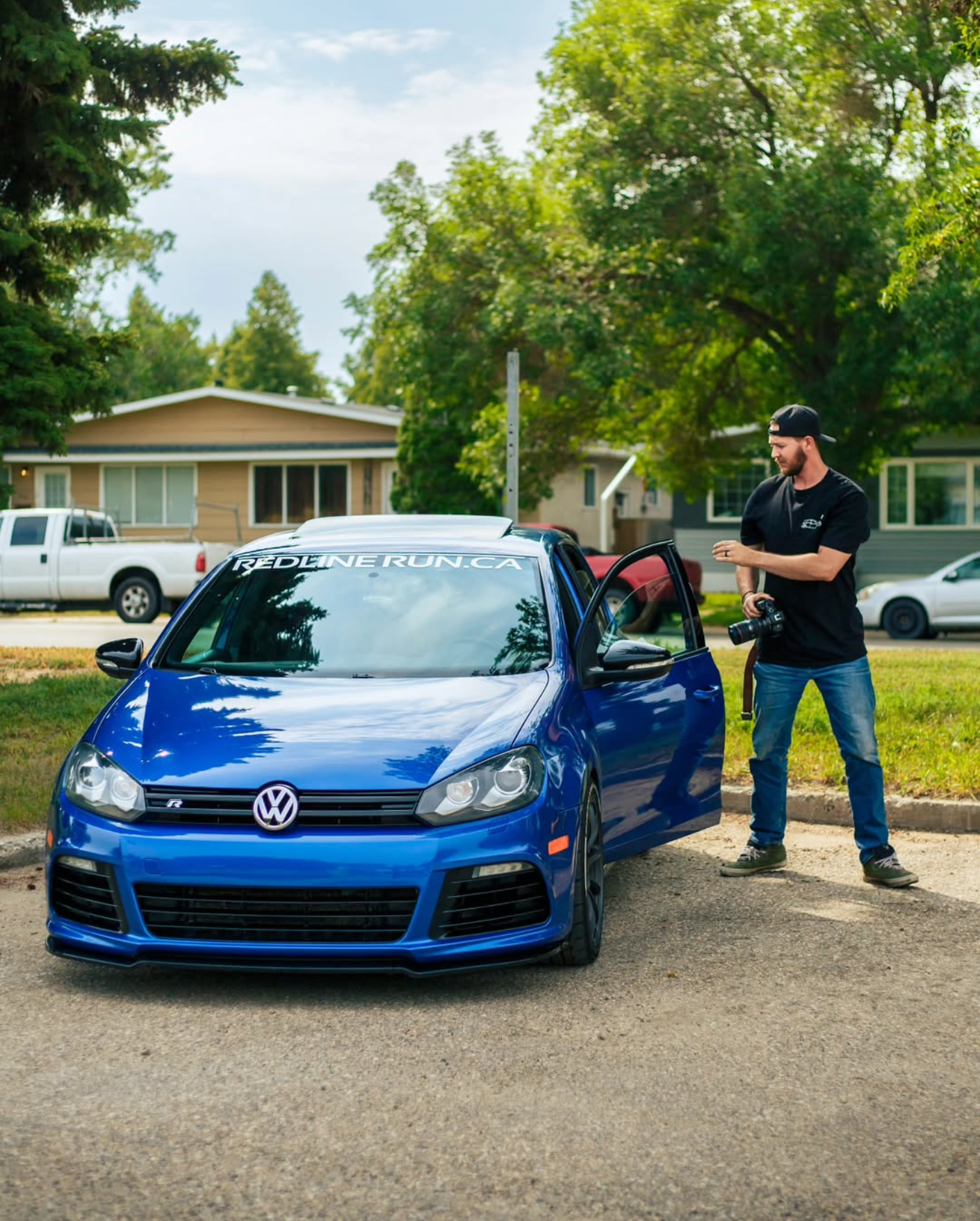 A man stands beside a blue Volkswagen car with an open door, holding a camera. The car has a window decal reading 'REDDINE.RUN.CA' and is parked on a residential street with trees and houses in the background.