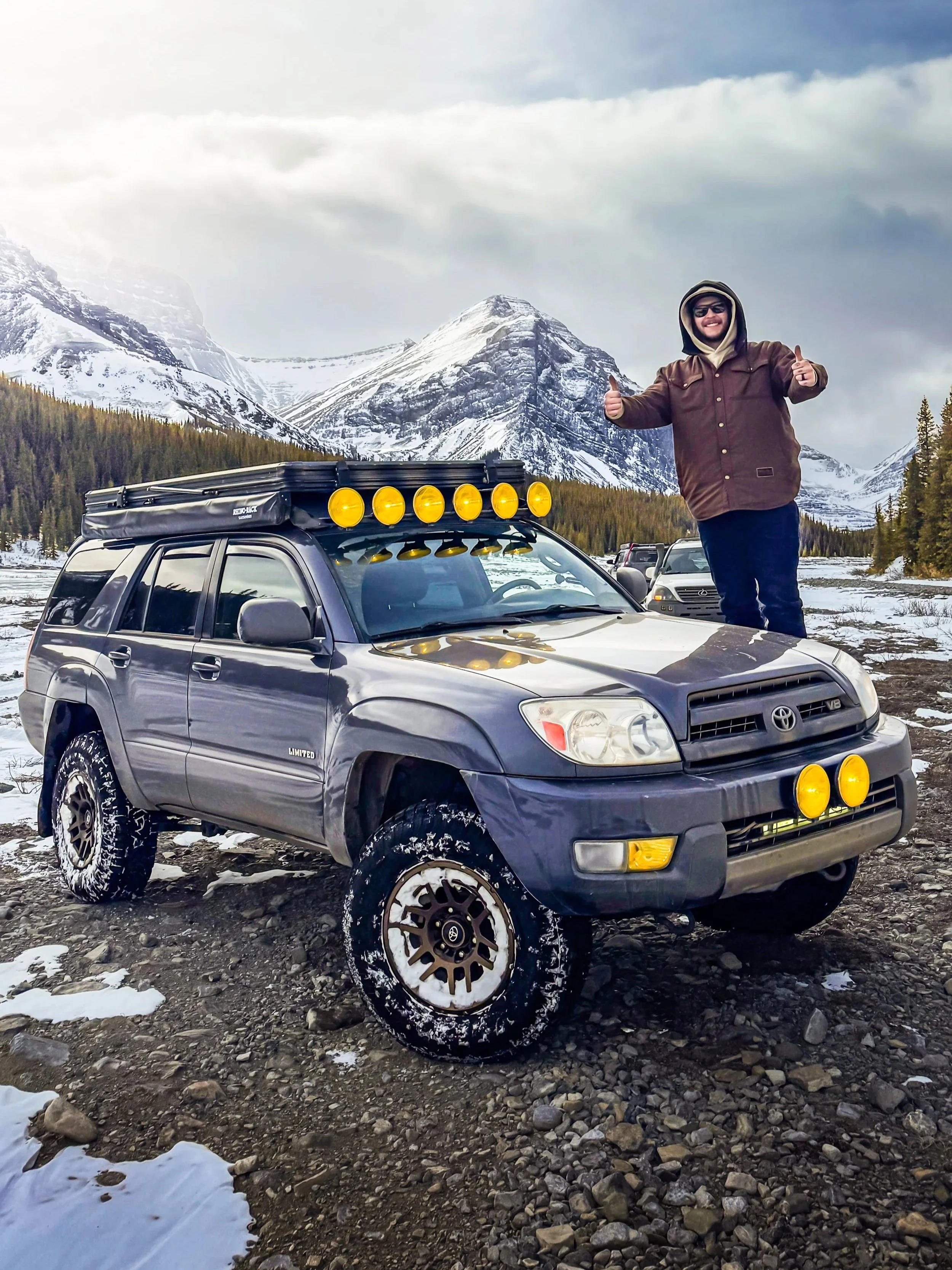 A smiling man in winter clothing standing next to a dark gray Toyota SUV with roof lights, in a snowy mountain landscape with pine trees and snow-capped peaks.