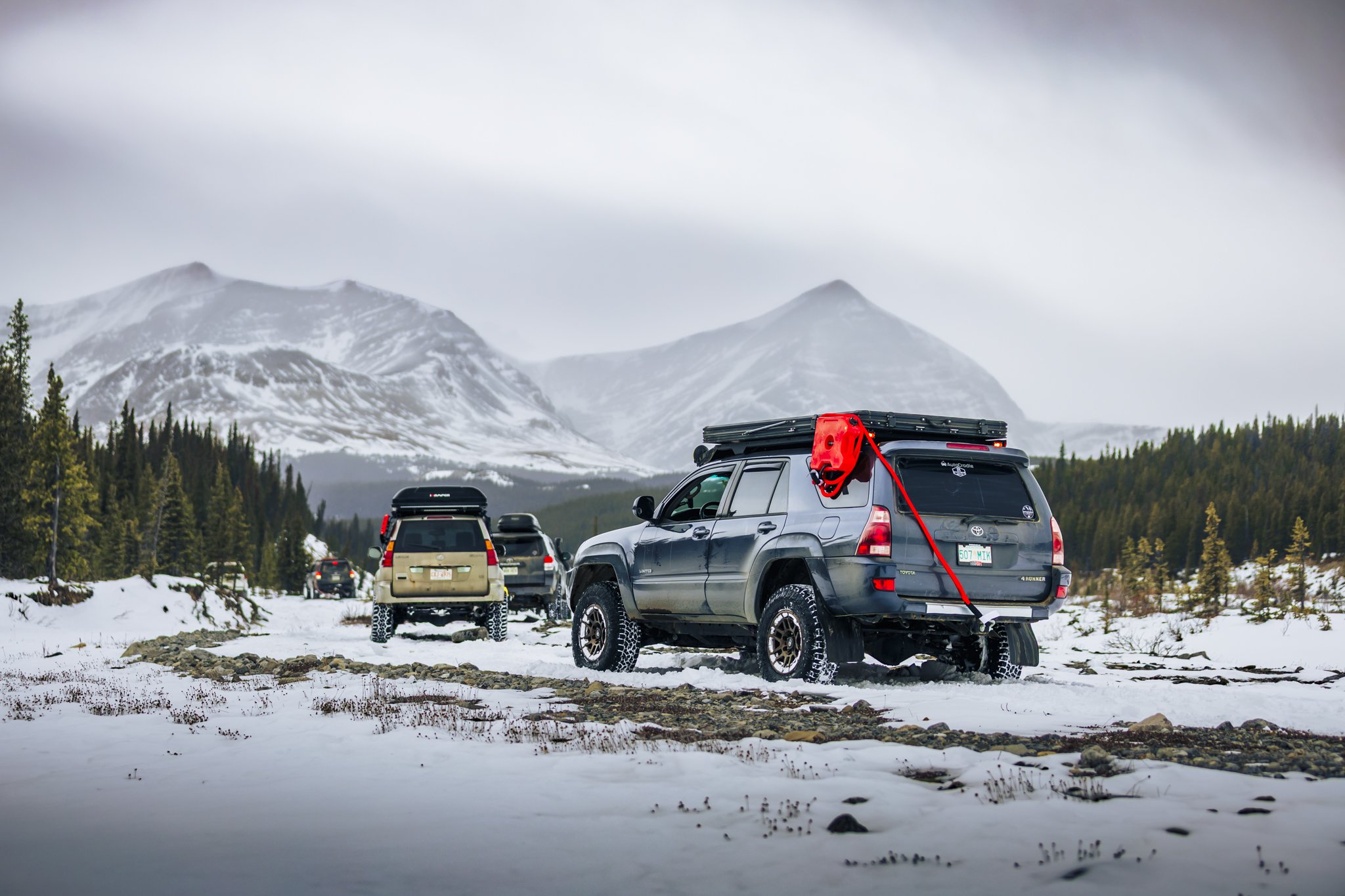Four vehicles are parked on a snowy mountain landscape, with snow-covered mountains and a cloudy sky in the background.