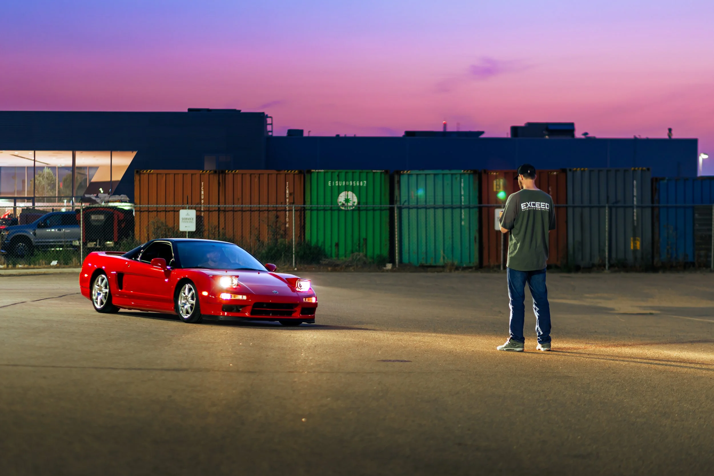 A red sports car parked on the empty lot during dusk, with a person standing nearby looking at their phone, and a background of colorful shipping containers and a modern building.